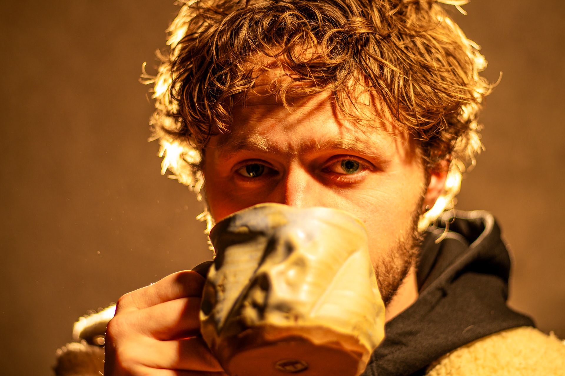A close-up of a man with curly hair and a beard, drinking from a unique, textured ceramic mug that covers the lower half of his face. His bright blue eyes look directly at the camera, and he is backlit by a warm, golden light.