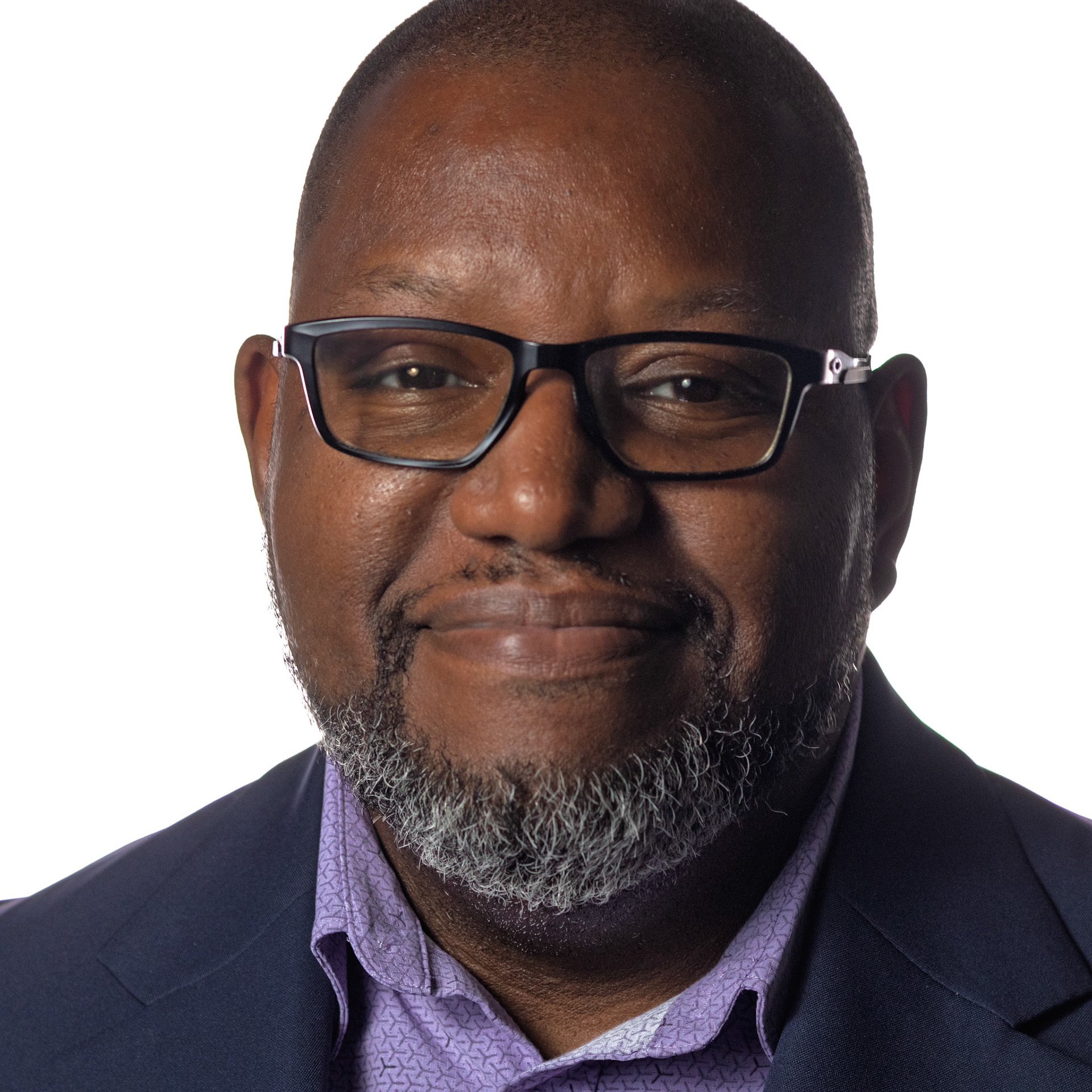 A professional headshot of a Black man with a salt-and-pepper beard, wearing glasses, a navy blazer, and a patterned purple shirt. He is smiling calmly at the camera in front of a white background.