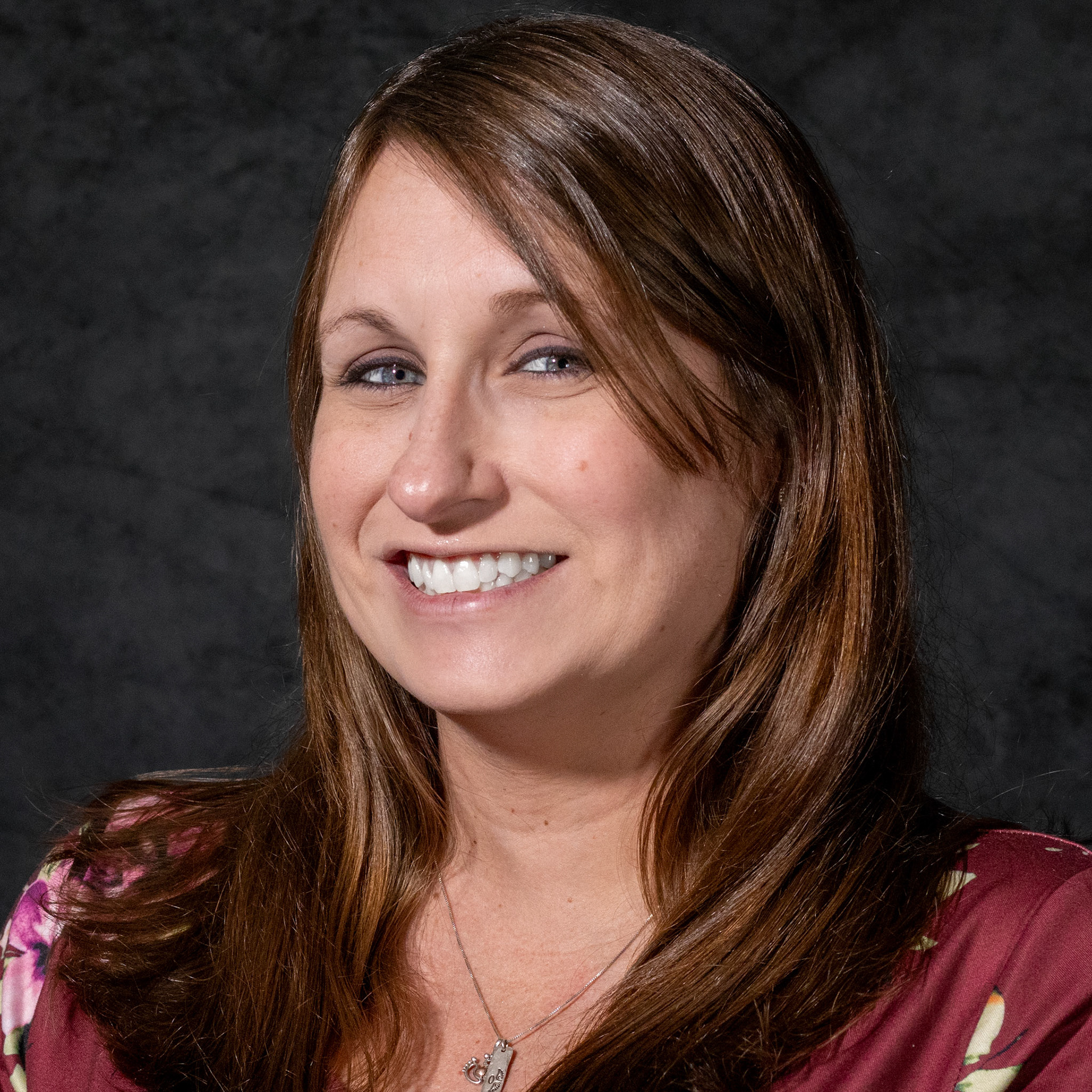 A professional headshot of a smiling woman with long brown hair. She is wearing a maroon floral top and a silver necklace with a heart-shaped charm. The background is a dark, mottled grey.