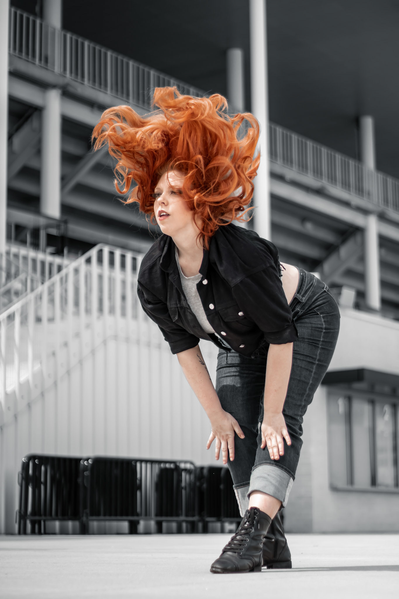 A woman with bright red, voluminous hair is bent over with her hands on her knees, looking to the side. She is wearing a black jacket, dark jeans, and black combat boots. The background is a grey stadium with white stairs and railings.