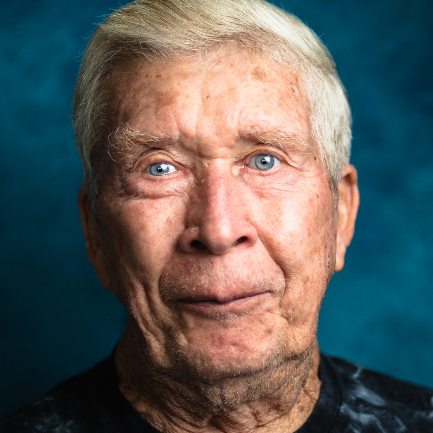 A close-up portrait of a senior man with white hair and bright blue eyes, looking directly at the camera with a slight smile. He is wearing a dark t-shirt against a dark blue mottled background.