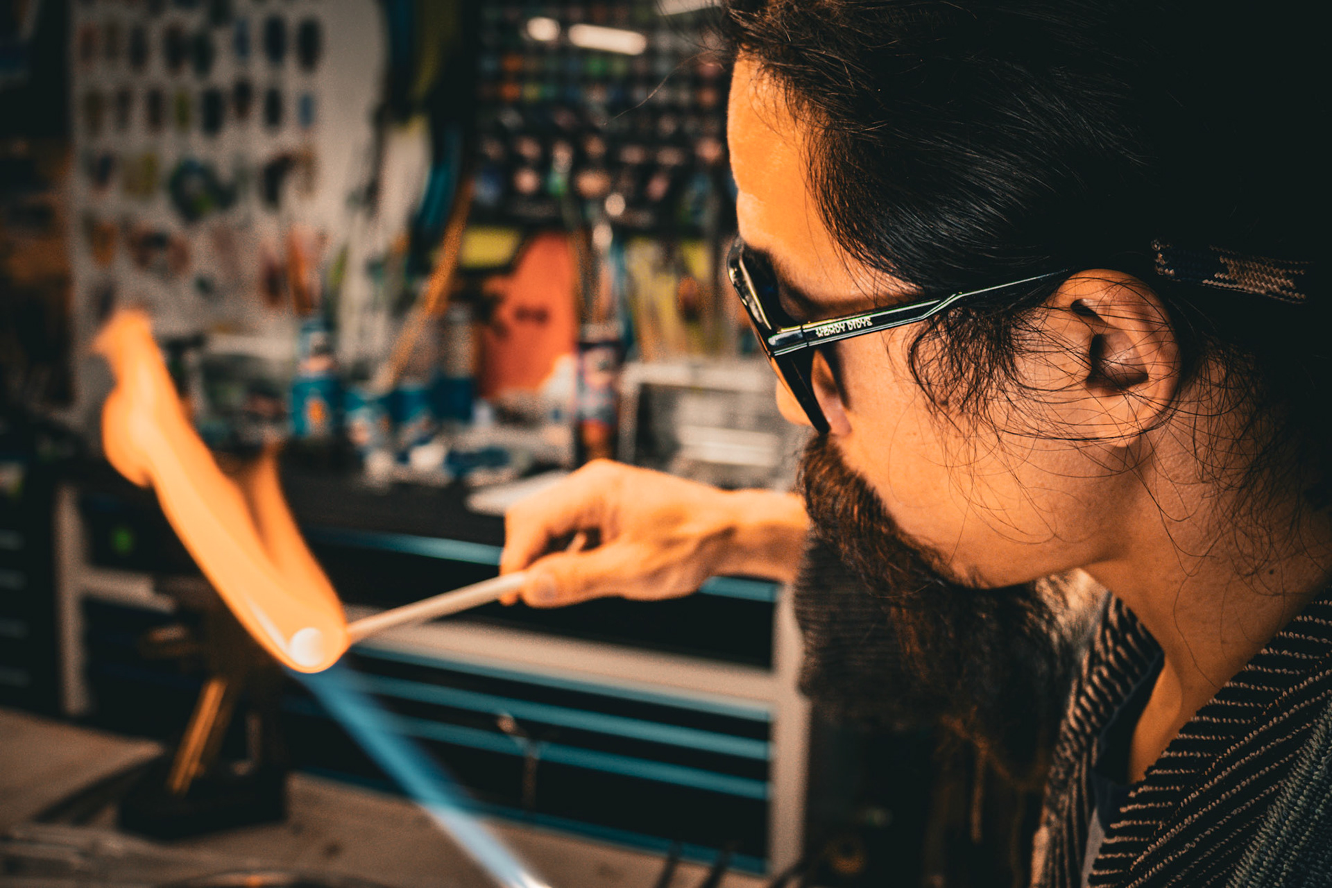 A close-up side profile of a man with long dark hair and a beard, wearing glasses, and leaning over a workbench. He is using a blowtorch to heat a clear glass rod, with a bright orange flame visible.