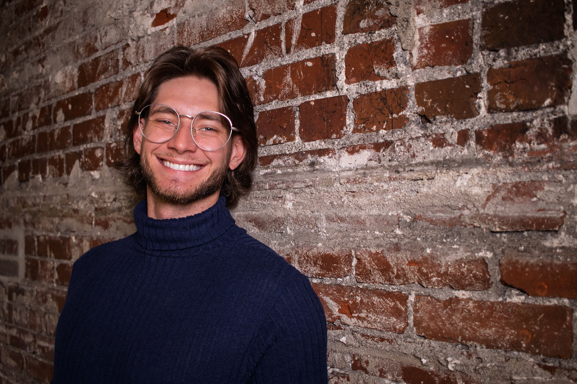 A smiling man with long brown hair, a beard, and round glasses, wearing a dark blue turtleneck sweater. He is posing for a headshot against an old, rustic brick wall.