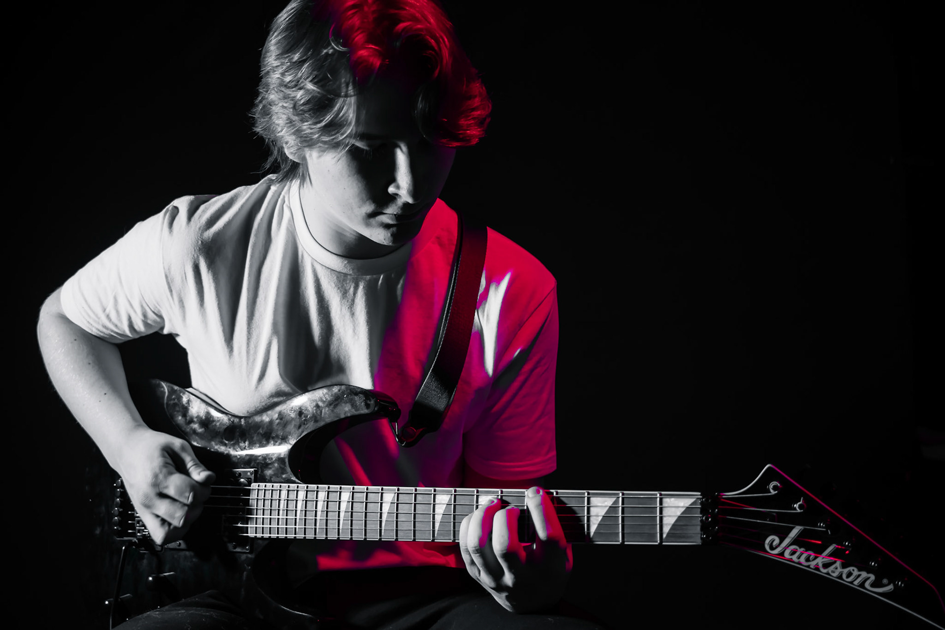 A young person with short, reddish hair plays an electric guitar. The image is mostly black and white, but a bright red light illuminates their hair, right shoulder, and a portion of the guitar. The background is completely black.