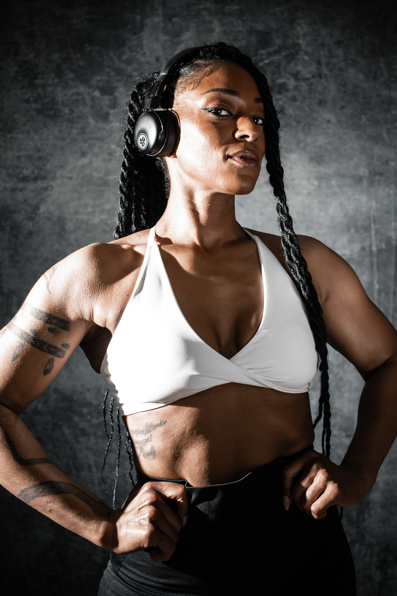 A strong Black woman with long braids, wearing headphones, a white sports bra, and black pants. She stands with her hands on her hips, looking directly at the camera with a confident expression. The background is a dark, mottled grey.