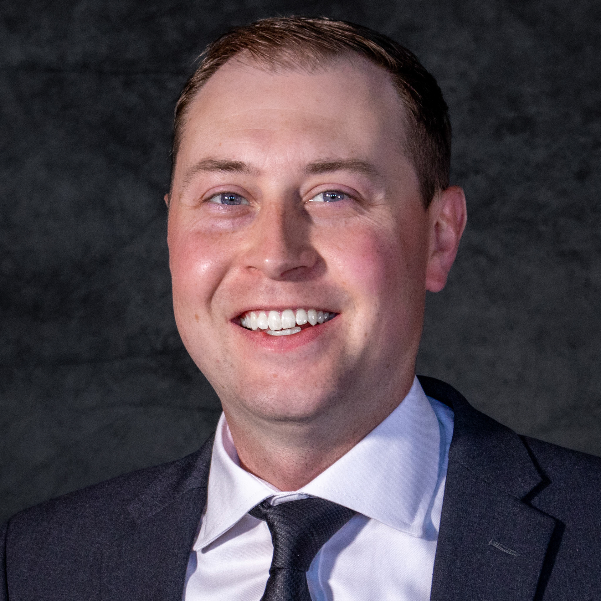 A professional headshot of a smiling man in a dark grey suit, white shirt, and black patterned tie. He is smiling widely, showing his teeth, against a dark, mottled grey background.