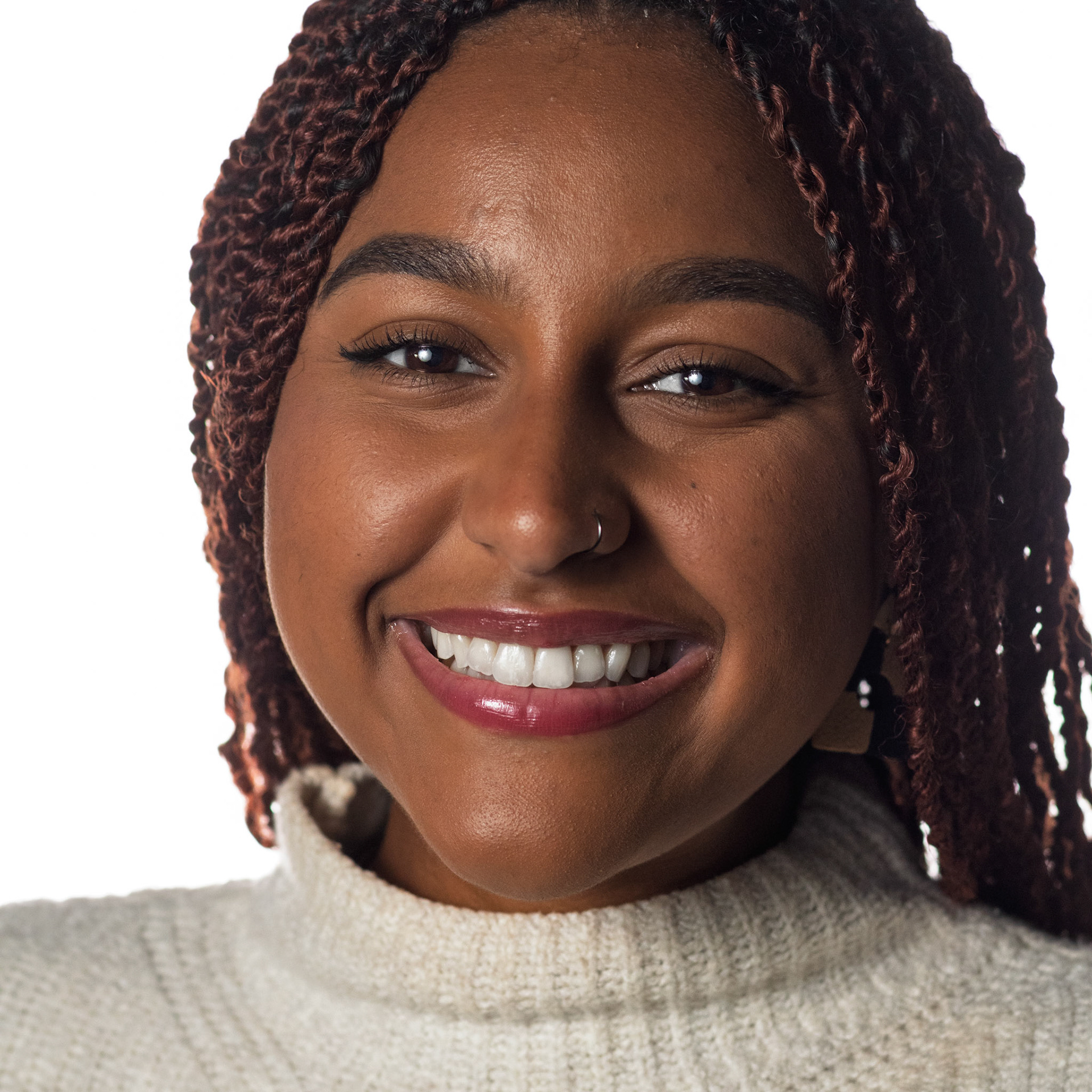 A close-up portrait of a young Black woman with reddish-brown braided hair. She is smiling broadly, showing her white teeth, and has a nose ring. She is wearing a cream-colored turtleneck sweater, and the background is plain white.
