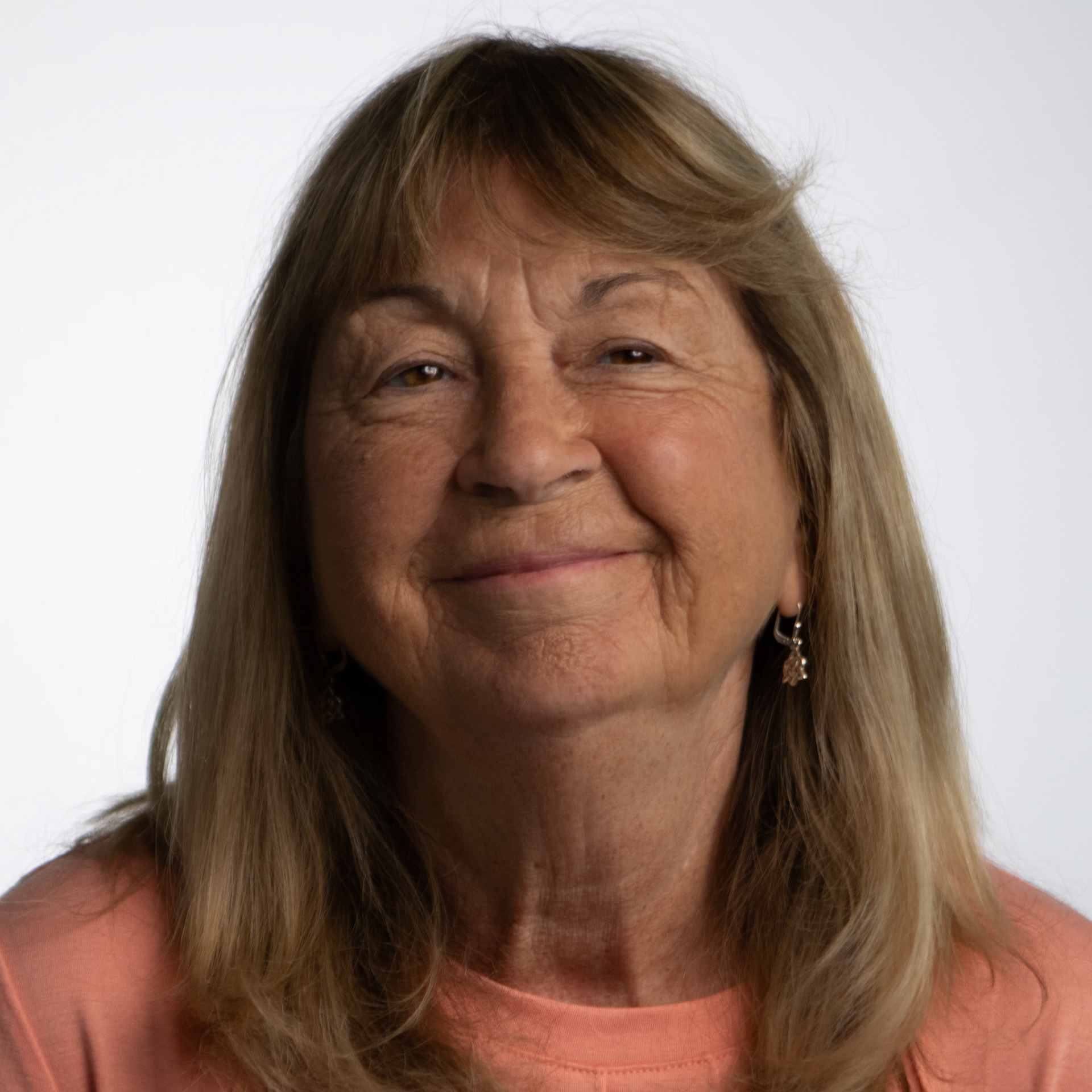 A headshot of an elderly woman with shoulder-length blond hair. She is smiling gently at the camera and wearing a peach-colored top against a plain white background.