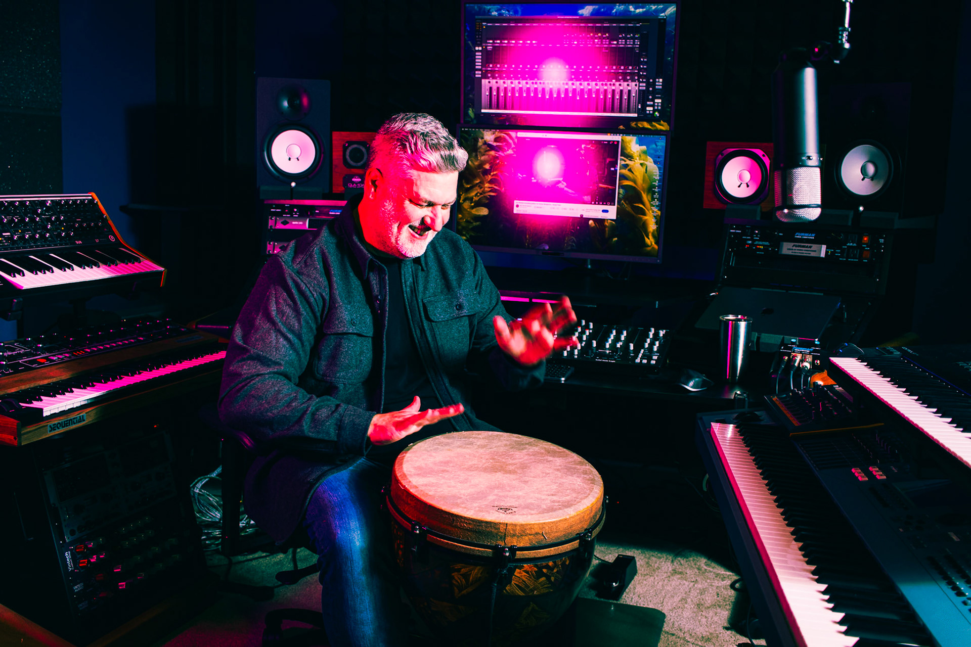A man with a grey beard, wearing a dark jacket, sits in a neon-lit music studio. He is playing a djembe drum with his hands, surrounded by keyboards, synthesizers, and computer monitors displaying sound wave patterns.