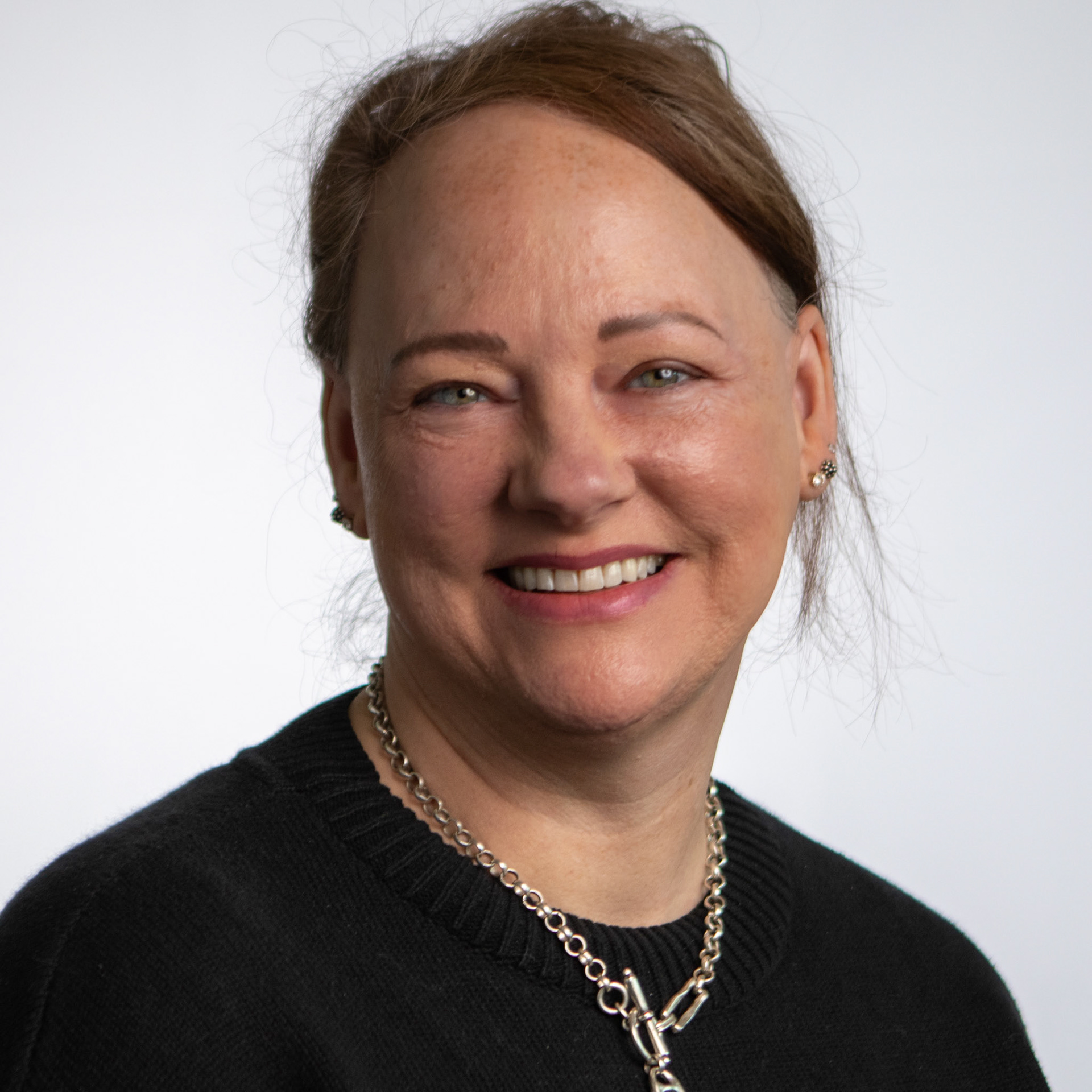 A headshot of a woman with light brown hair, smiling with her teeth showing. She is wearing a black sweater and a chunky silver chain necklace. The background is a plain white studio wall.