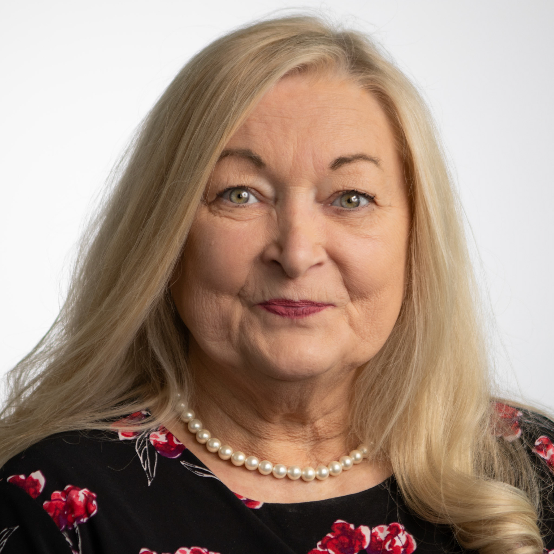 A headshot of an elderly woman with long blonde hair and a pearl necklace. She is wearing a black shirt with a red and white floral pattern and looking directly at the camera. The background is a plain white studio wall.
