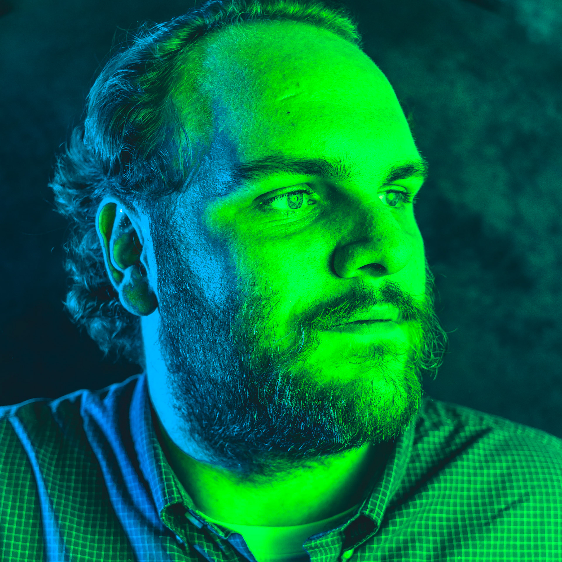 A portrait of a man with a beard, looking off to the side. The image is lit with a split-color effect, with the left side of his face and shoulder illuminated in blue and the right side of his face in a bright green.