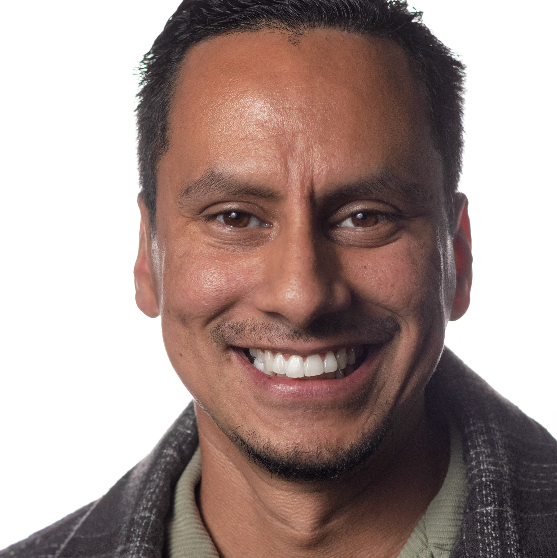 A headshot of a Hispanic man with dark hair and a small goatee. He is smiling widely, showing his teeth, and wearing a grey patterned jacket over a light green shirt. The background is white.