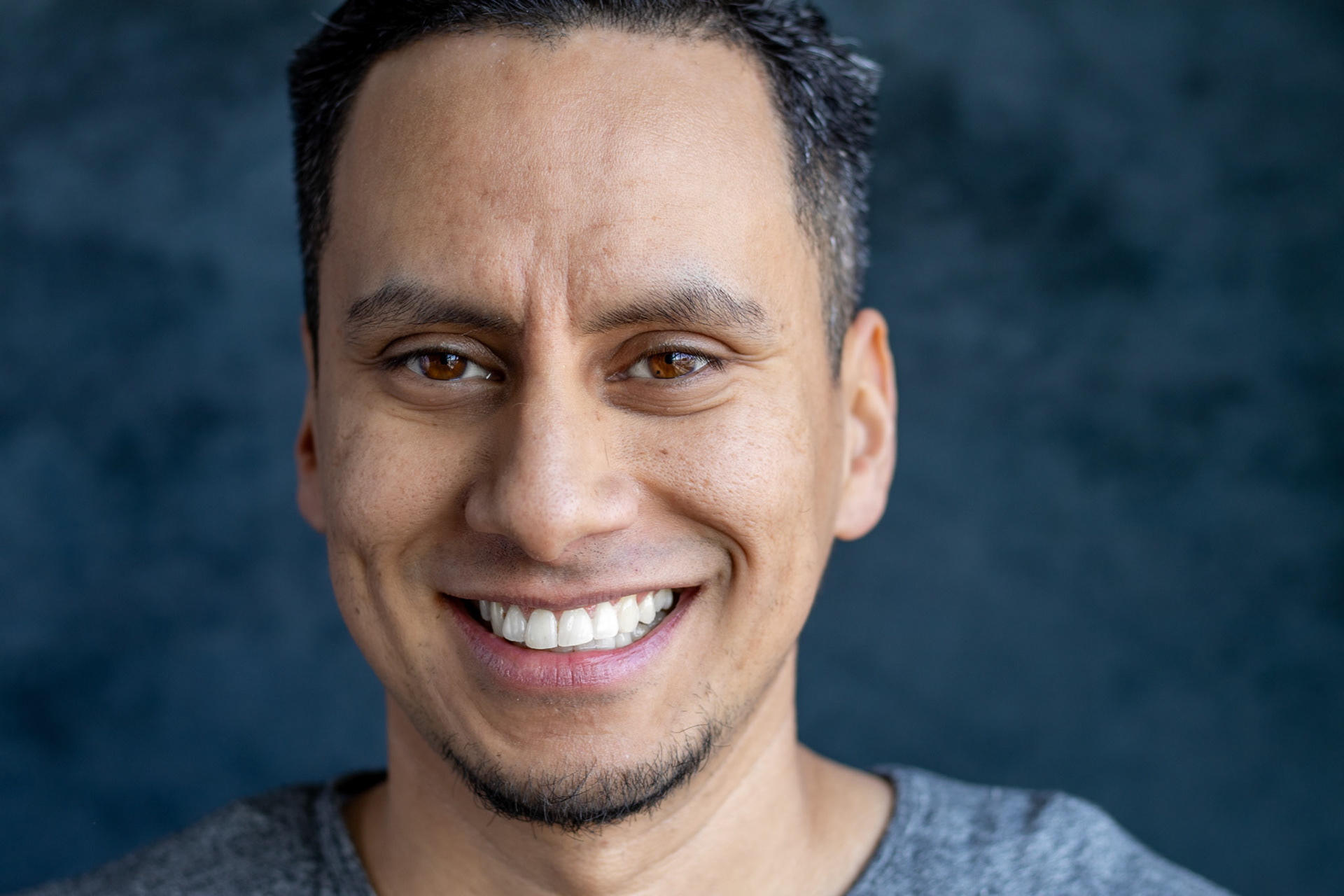 A close-up portrait of a smiling man with short hair and a small goatee. He is wearing a grey t-shirt and smiling broadly at the camera.