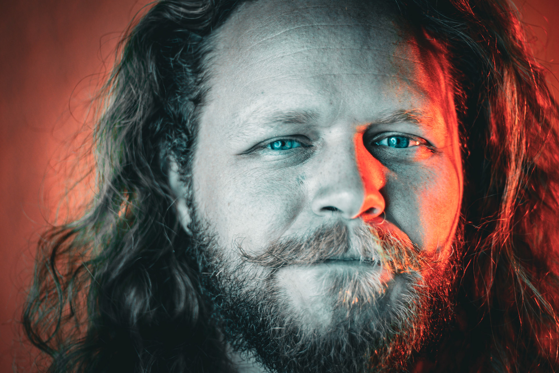 A close-up portrait of a man with long hair and a beard. His face is lit with a dramatic split-lighting effect, with a red glow on the right side and a desaturated, cool tone on the left. His intense blue eyes look directly at the camera.