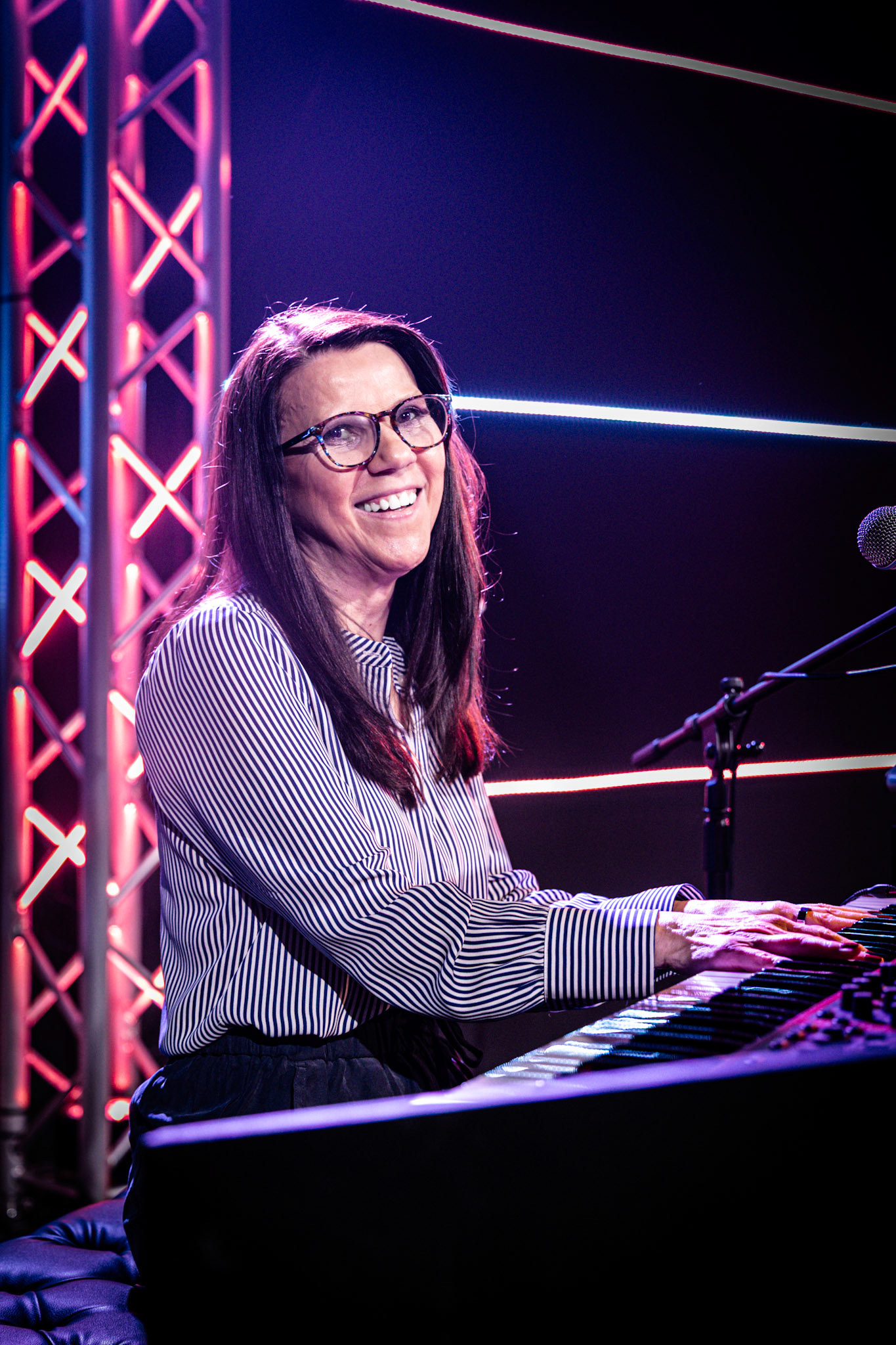 A smiling woman with long brown hair and glasses plays a keyboard on a dimly lit stage. She is wearing a striped black and white long-sleeve shirt. The background is dark, with pink and blue neon lights.