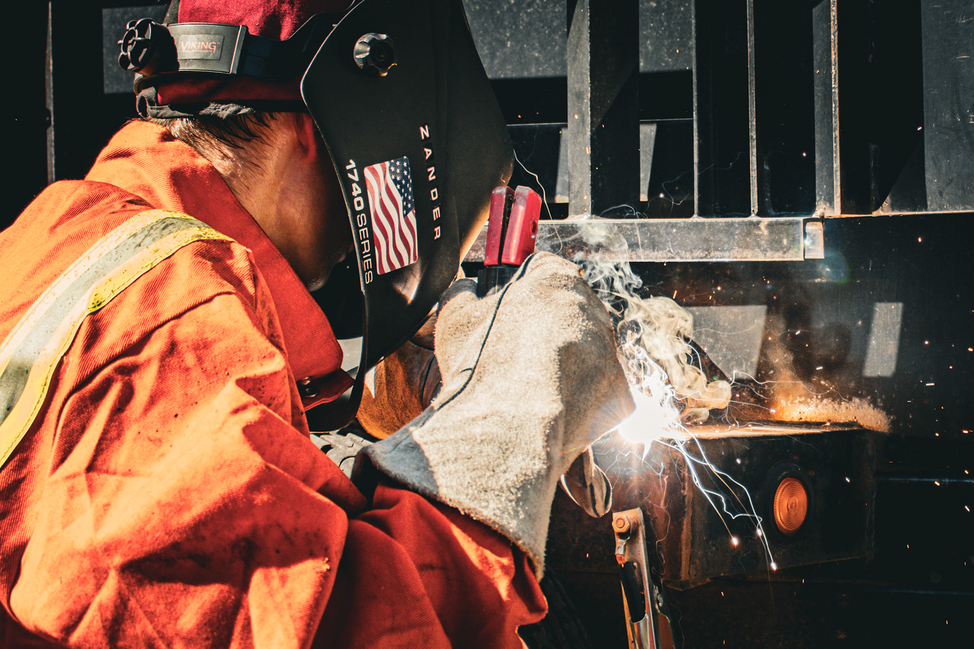 A welder wearing a helmet with an American flag sticker is working on a metal structure. Sparks and light fly from the welding torch, and he is wearing a red jacket and white gloves.