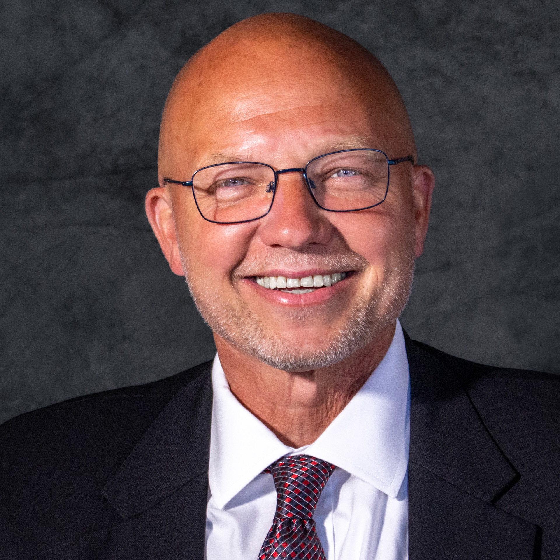 A professional headshot of a bald man with a goatee and glasses, smiling broadly at the camera. He is wearing a black suit jacket, a white shirt, and a red and black striped tie. The background is a dark, mottled grey.