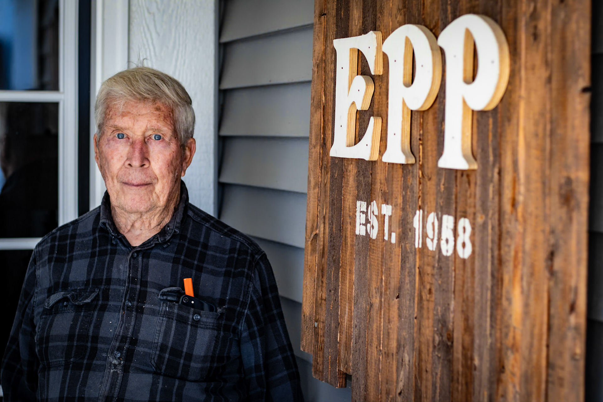 A waist-up shot of a senior man with white hair and bright blue eyes, wearing a grey plaid button-down shirt. He is standing in front of a house, next to a large wooden sign that reads "EPP EST. 1958."