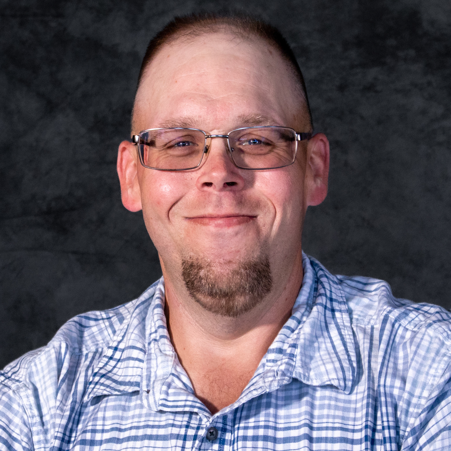 A headshot of a man with a shaved head, a goatee, and glasses. He is smiling widely, showing his teeth, and wearing a blue and white plaid shirt. The background is dark and mottled.