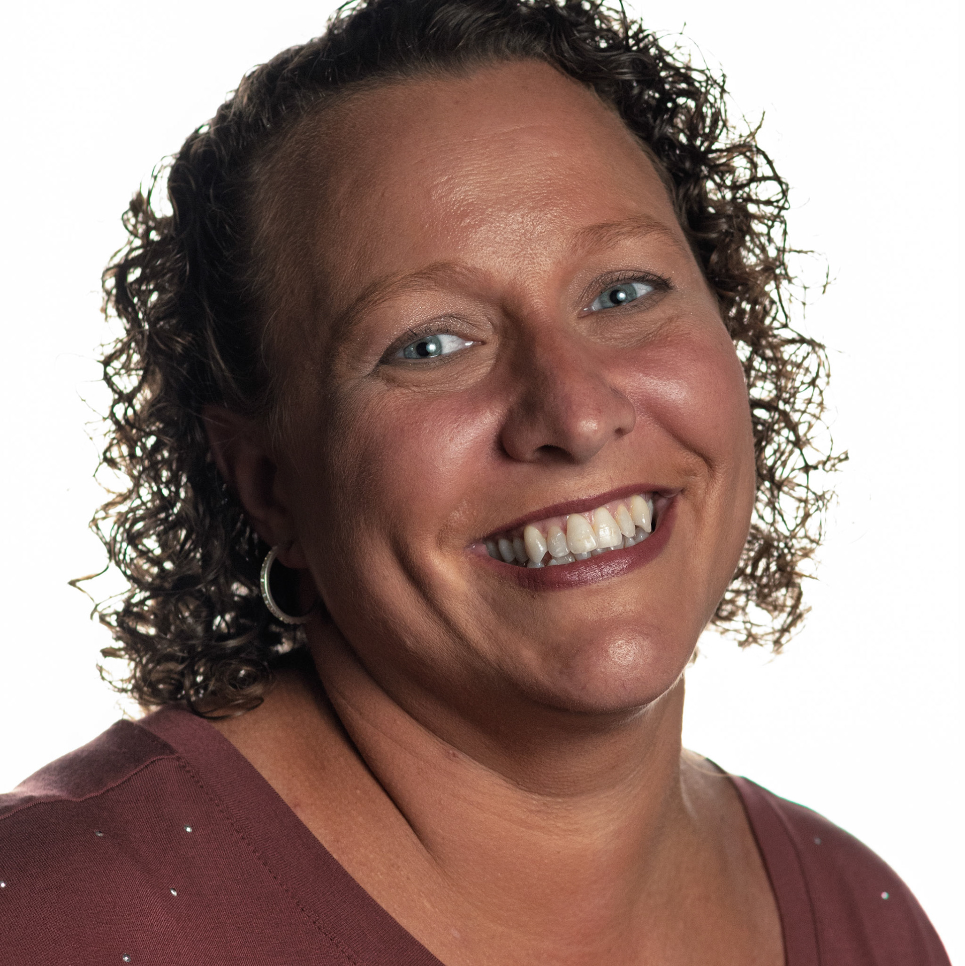 A headshot of a smiling woman with curly brown hair and bright blue eyes. She is wearing a reddish-brown shirt with small sparkly accents and a silver hoop earring. The background is white.
