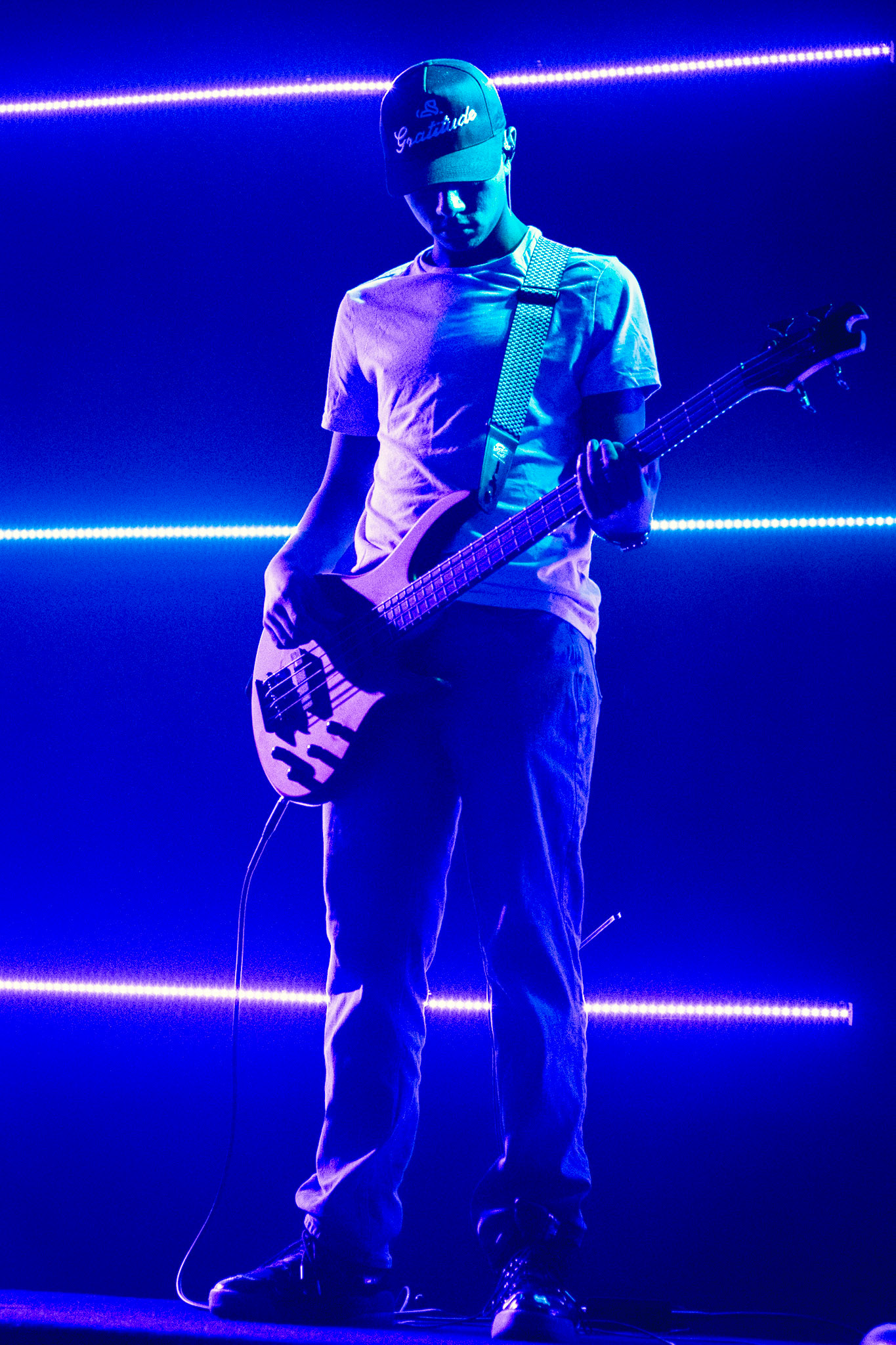 A full-body shot of a young person in a white t-shirt, jeans, and a baseball cap, playing a bass guitar on a dark stage. They are lit from above and below by bright blue neon lights.