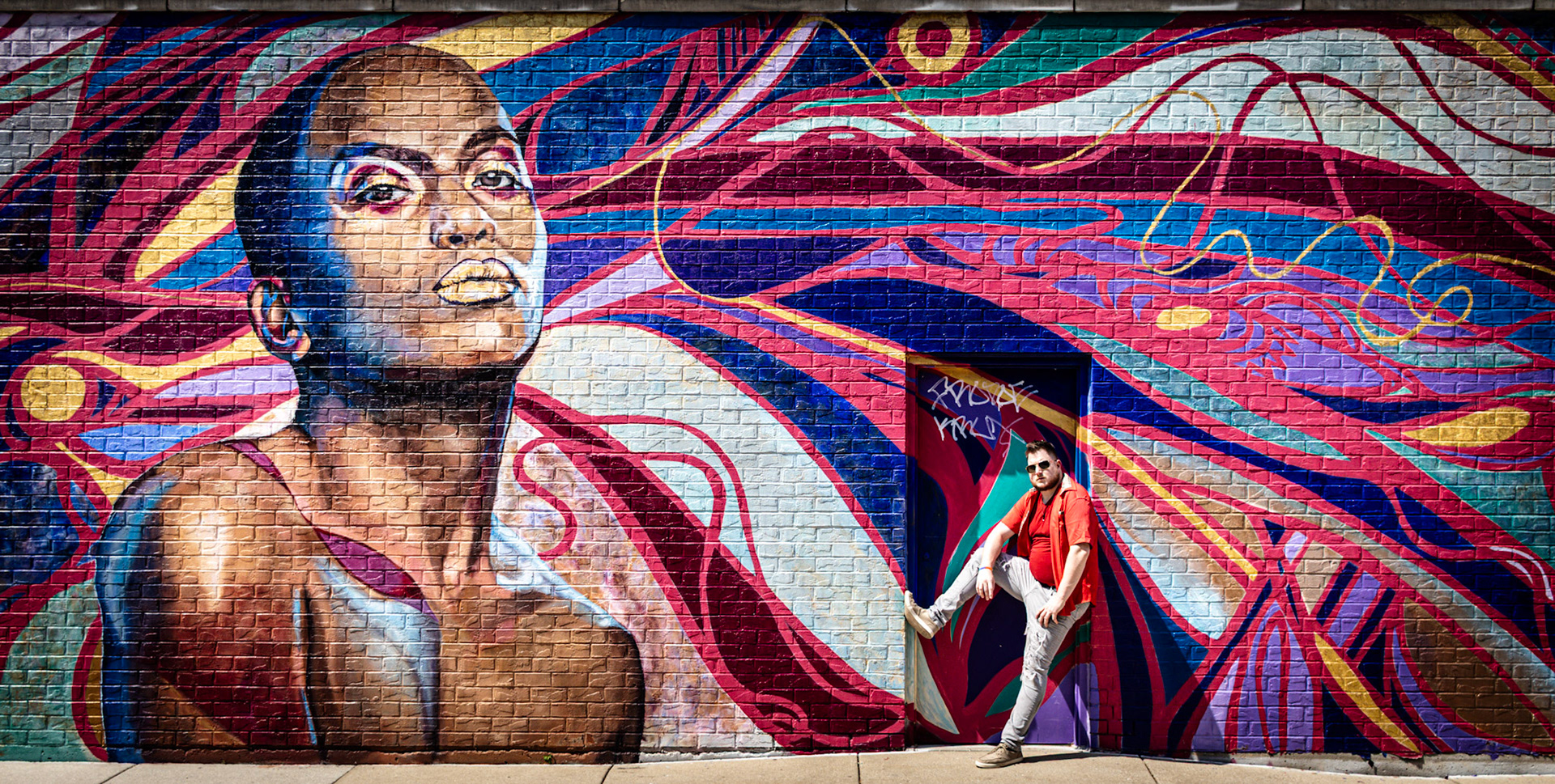 A man in a red shirt and sunglasses poses in front of a colorful mural on a brick wall. The mural features a large portrait of a person with gold lips and elaborate, abstract patterns in shades of red, blue, and purple.