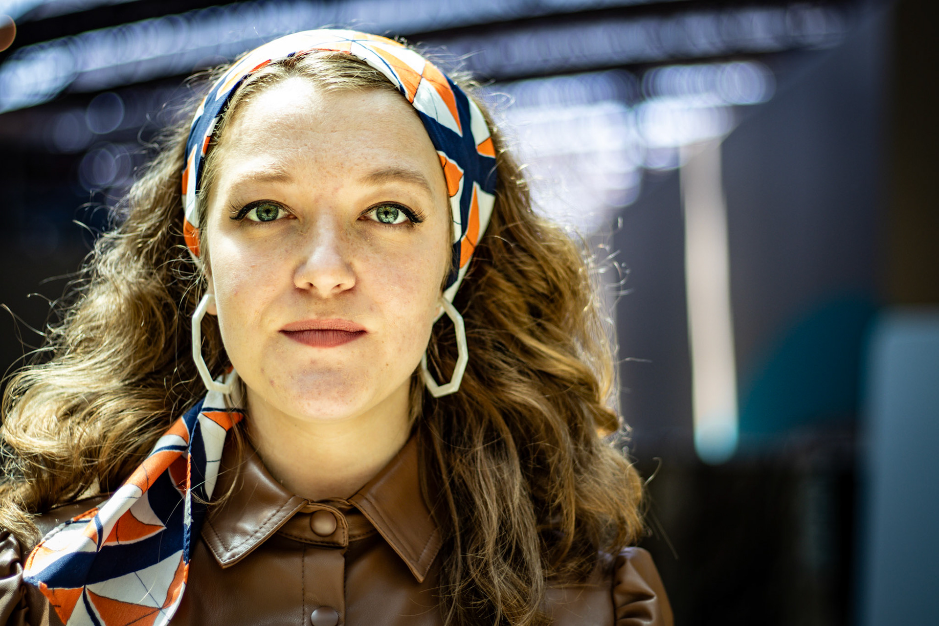 A portrait of a woman with long, curly brown hair and green eyes. She wears a colorful headscarf, large white geometric earrings, and a brown leather-like shirt. She is looking directly at the camera with a serious expression.