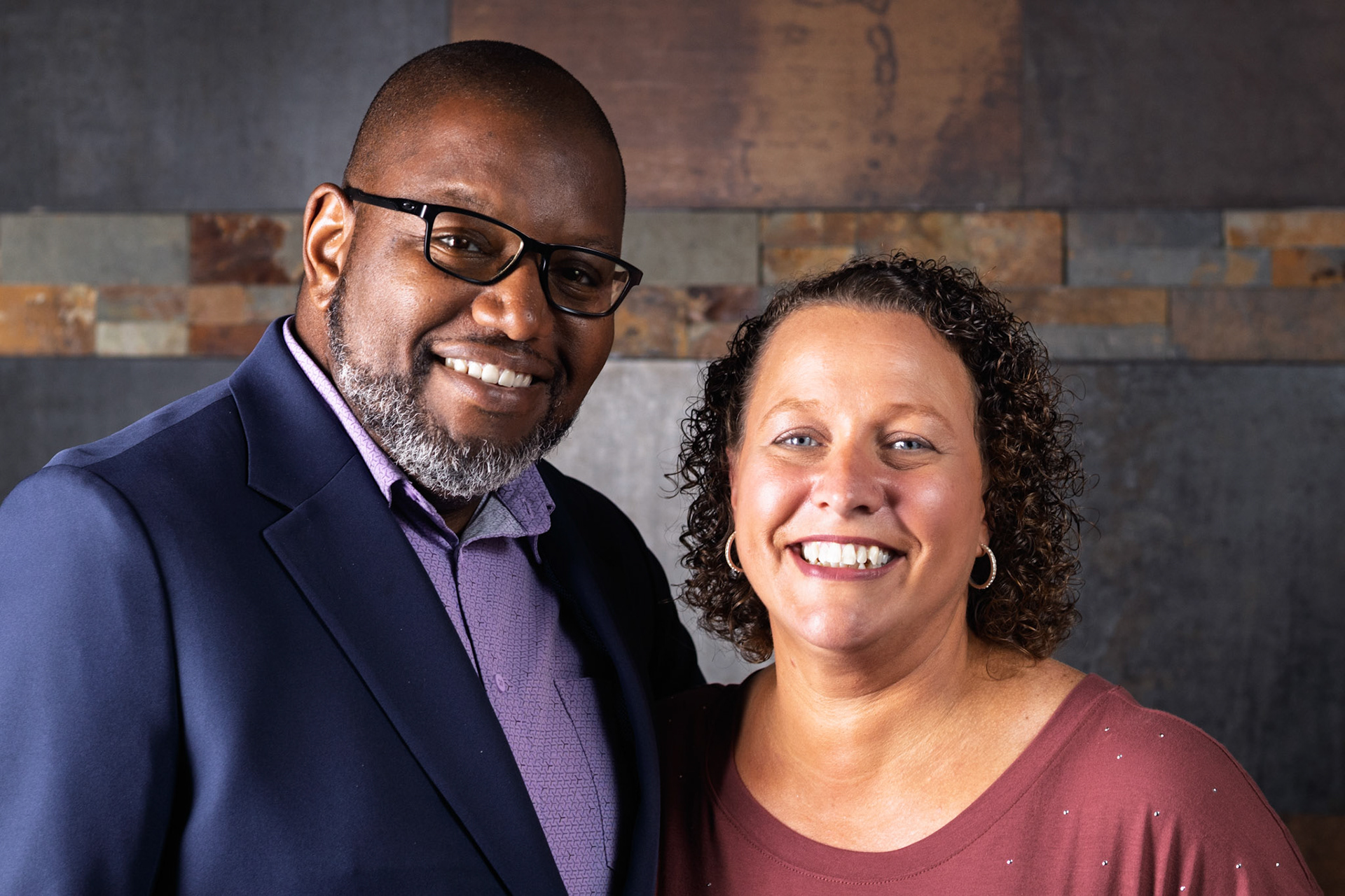A headshot of a Black man and a white woman smiling at the camera. The man wears a navy blazer and purple shirt, the woman a rusty red top. They stand close together against a stone wall background.