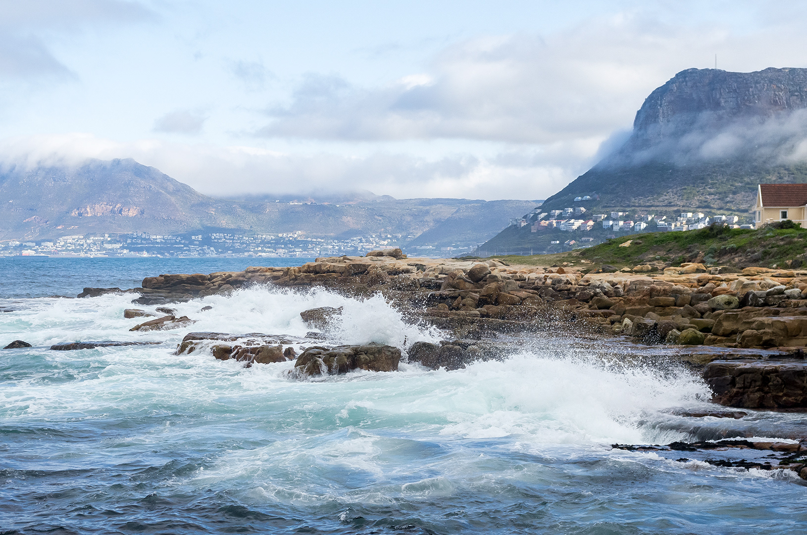 White top waves rusing over a rock waterfront.  Houses along the Cape coastline recede into the humid distance