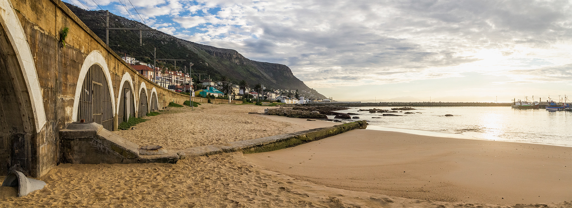 The white arches of the metroline curve into the distance of Kalk Bay Jetty.  The mountains descending into the waters with the bright morning sun just behind clouds