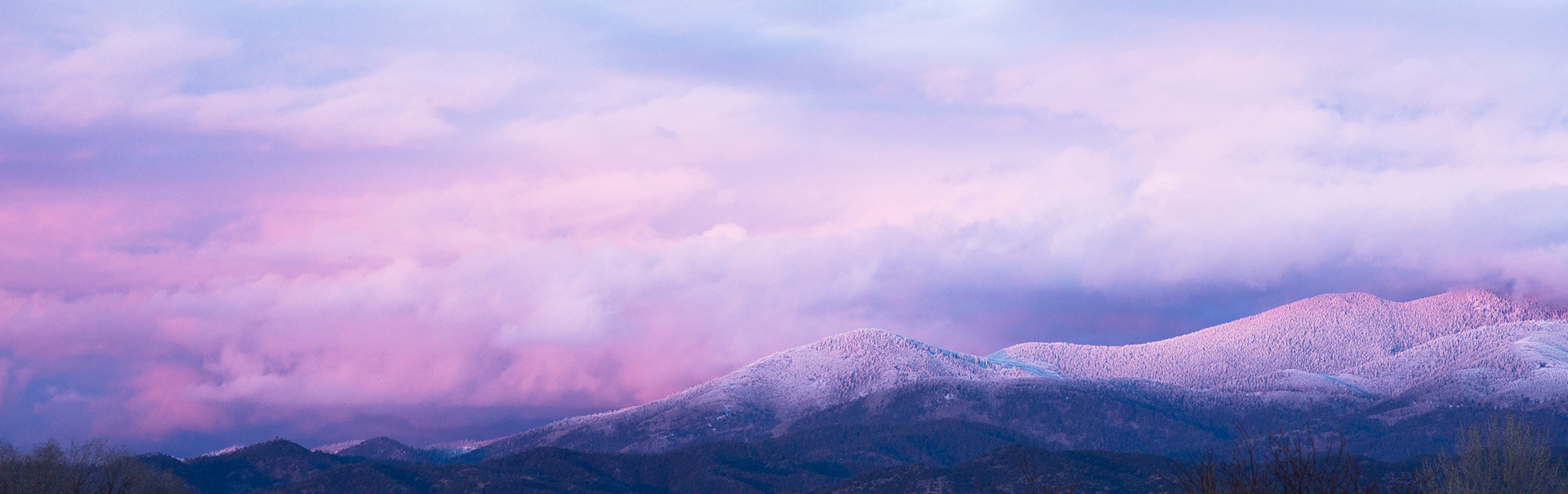 The white peaks of the Santa Fe National Forest rise above a dark winter treeline the clouds and snow glow salmon pink with sunset