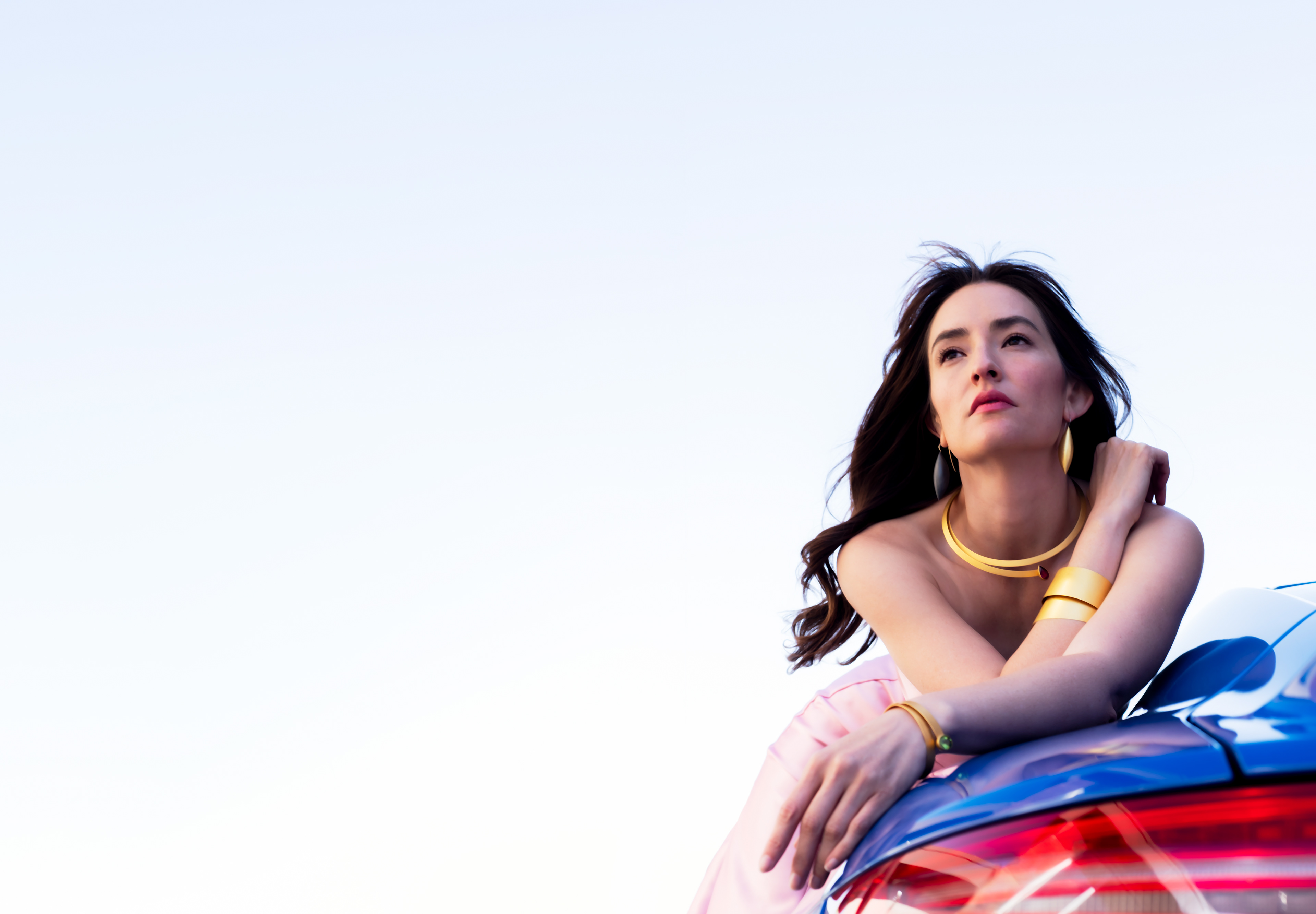 Jocelyn Montoya gazing pensively out to the distance horizon, a hazy white sky. She leans along the stylish lines of a blue Porsche 911, the red taillights enriching the flow of opulent hand hammered yellow jewelry of Ulla and Martin Kaufmann.