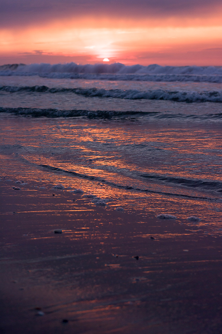 An orange and pink stormy morning with the sun just coming over the clouds and waves and reflecting in the lapping wave covered sand