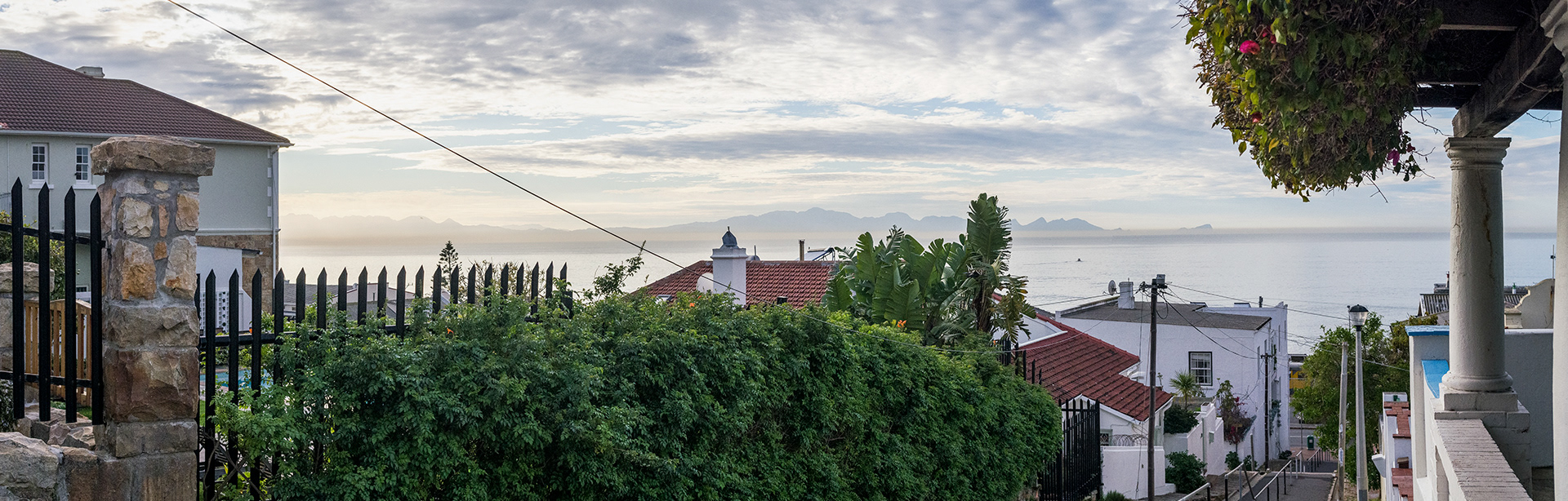 A view of False Bay and the mountains beyond from a vantage point over the rooflines, framed by a trellis column and a flowering bougainvellia