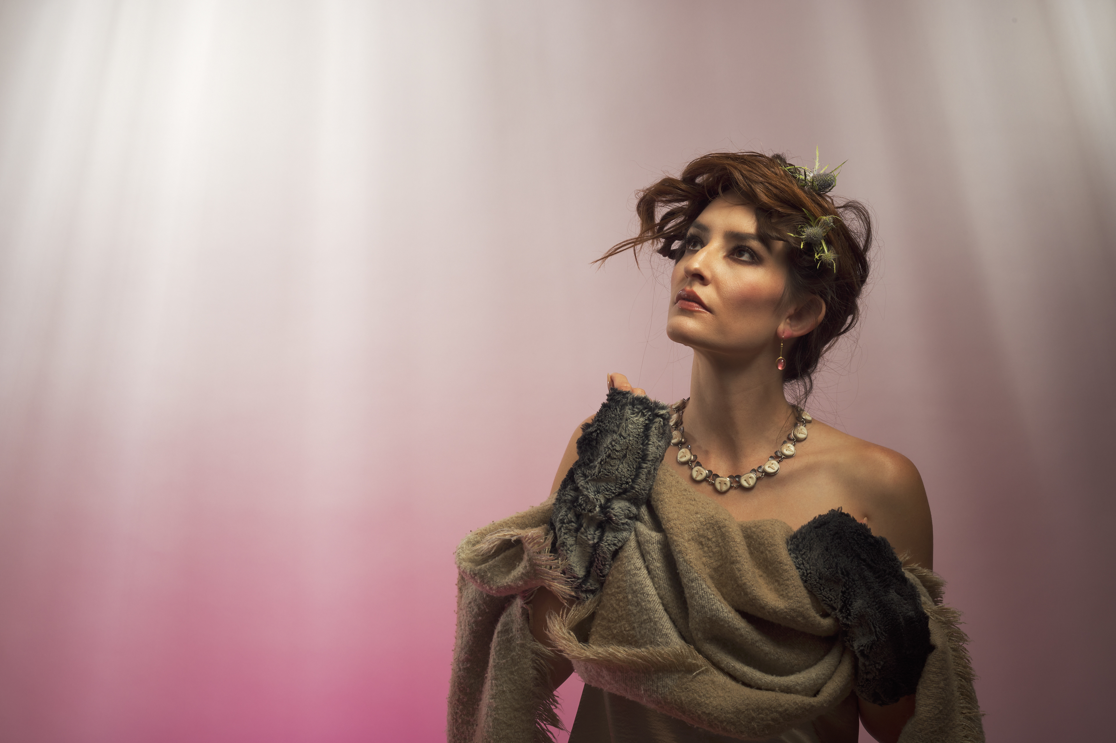 Jocelyn Montoya her gaze inquisitively upwards, is styled to look of the forest, with faux fur and dried thistles in her hair.  In the background white rays of light shine down and fade into a rusty pink mist.  She is wearing earrings and a necklace by Carolyn Morris Bach.