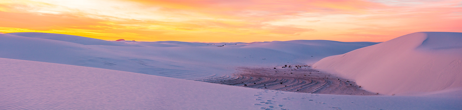 A cloudy sky of brilliant orange and salmon pink tinting the slope of a dune facing out towards an expanse of white sand to the horizon