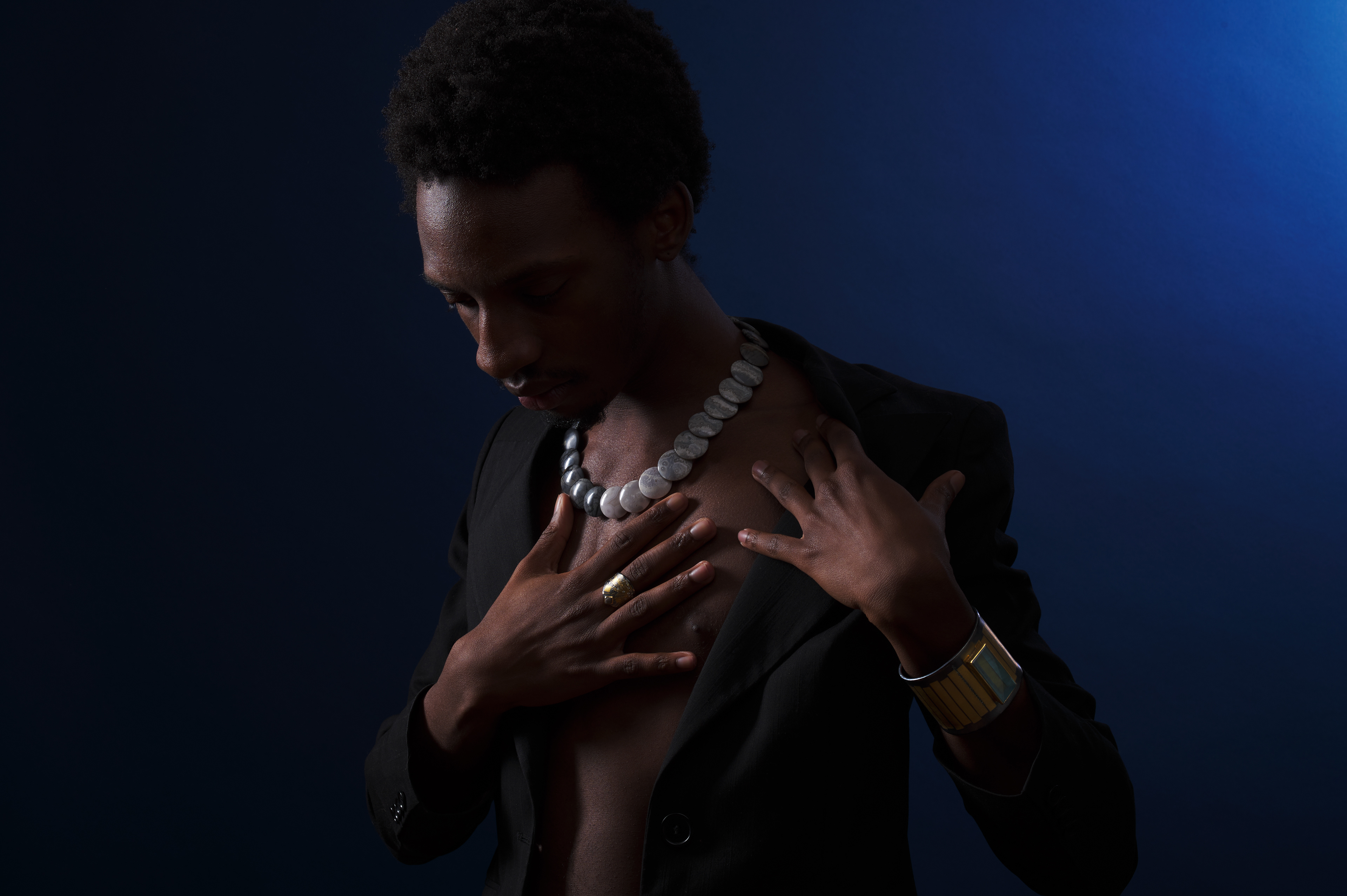 Jayson Lee, his gaze turned downward, shines in the blue darkness. The Atelier Zobel necklace of polished grey and white stones rests upon his touch, one gold and platinum ring draws your gaze.