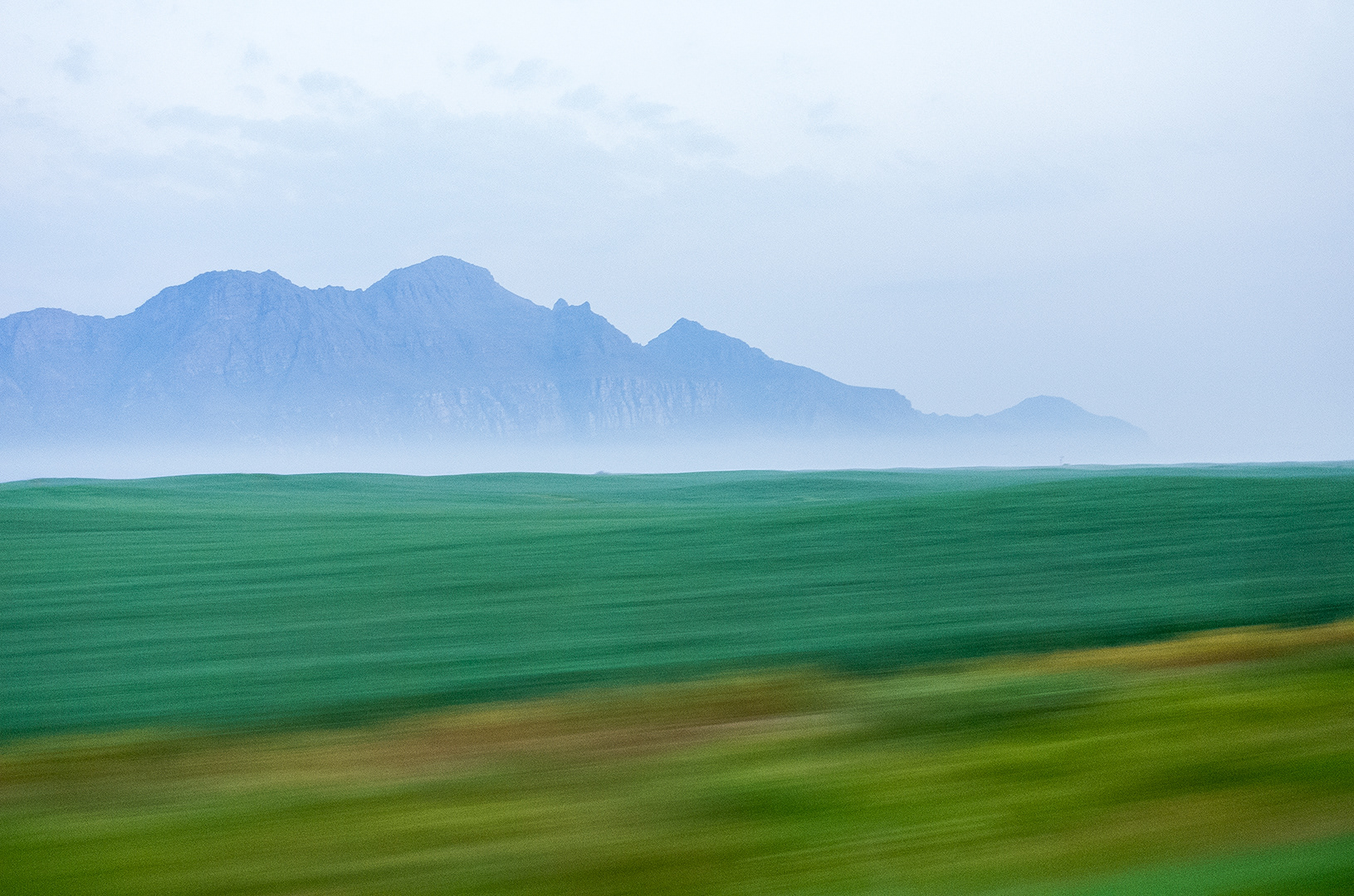 A long exposure from the car leaves the distant mountains sharp while the verdent green grasses near the road blur by, morning fog seperates the two