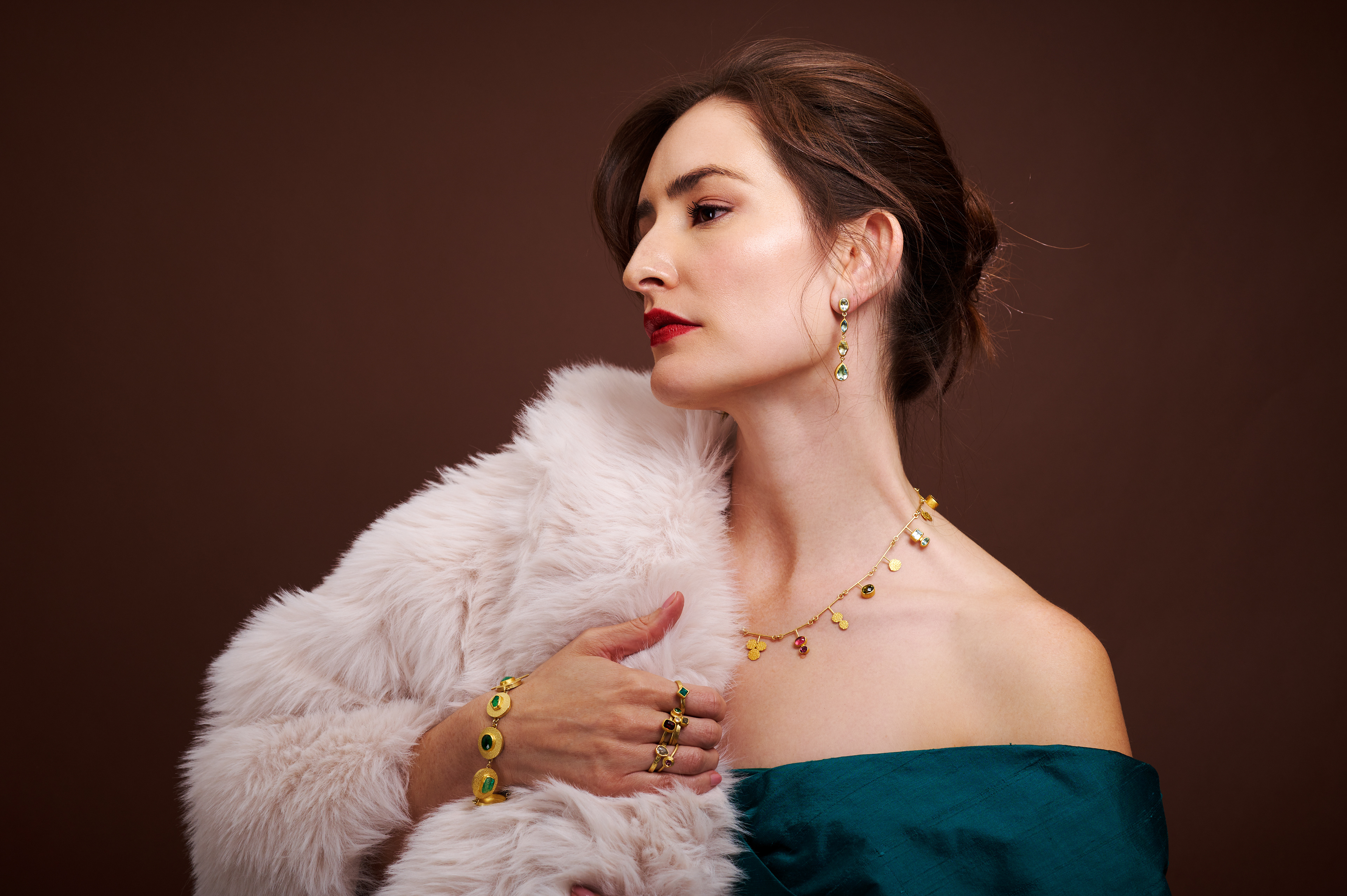 Jocelyn Montoya in a blue teal evening gown draped in a sumptuous white faux fur wearing a medley of yellow gold jewelry with a rainbow of stones by jeweler Petra Class, modern elegance..