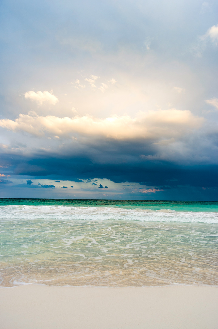 A soft aqua wave lapping on a white sand beach beneath a dark suntopped cloud.