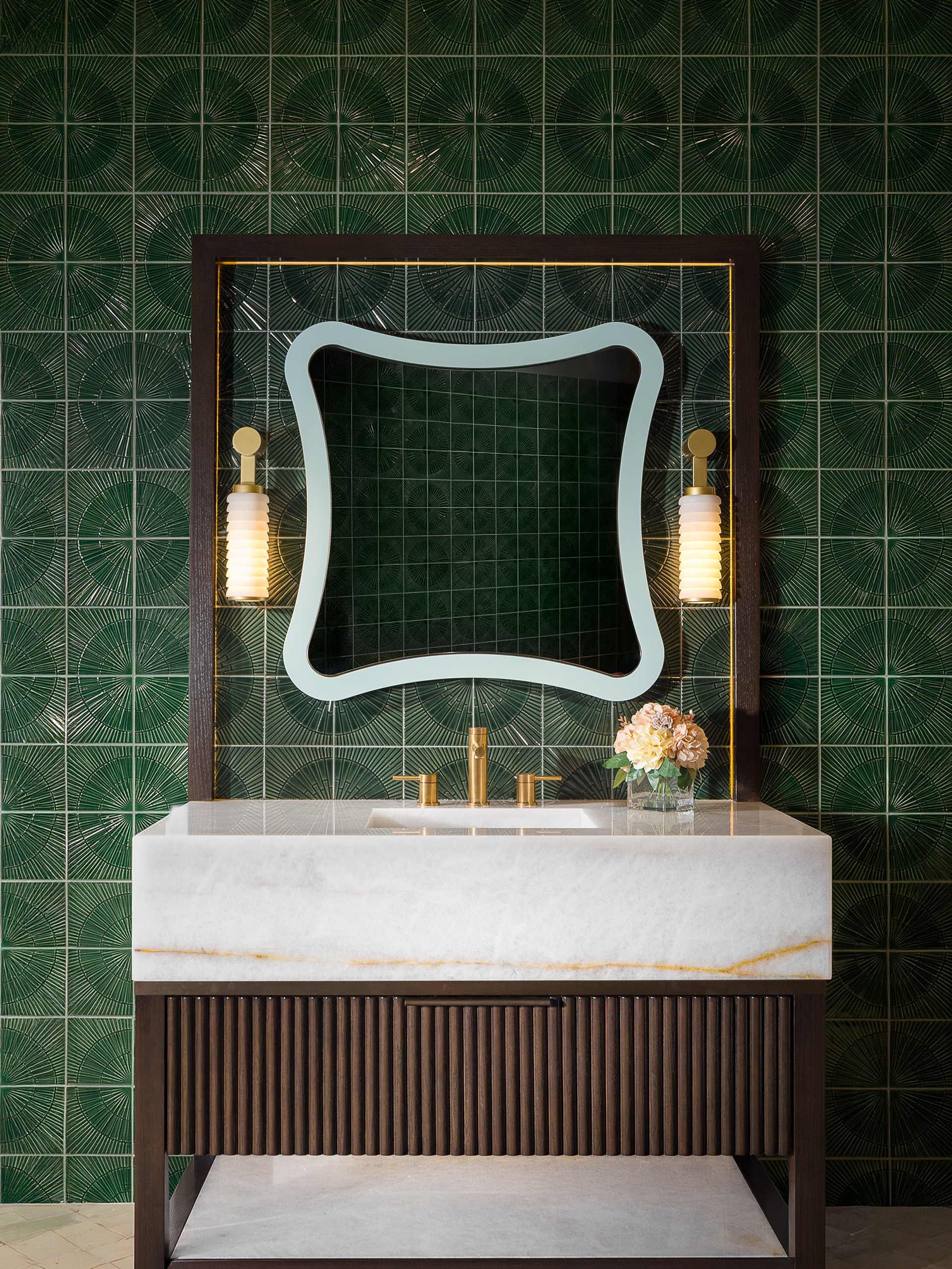 Architectural interior photography of a luxury powder room with emerald green geometric tile walls, curved statement mirror, brass sconces, marble vanity, and dark walnut cabinetry. Captured with balanced natural lighting and centered composition for hospitality and interior design marketing.