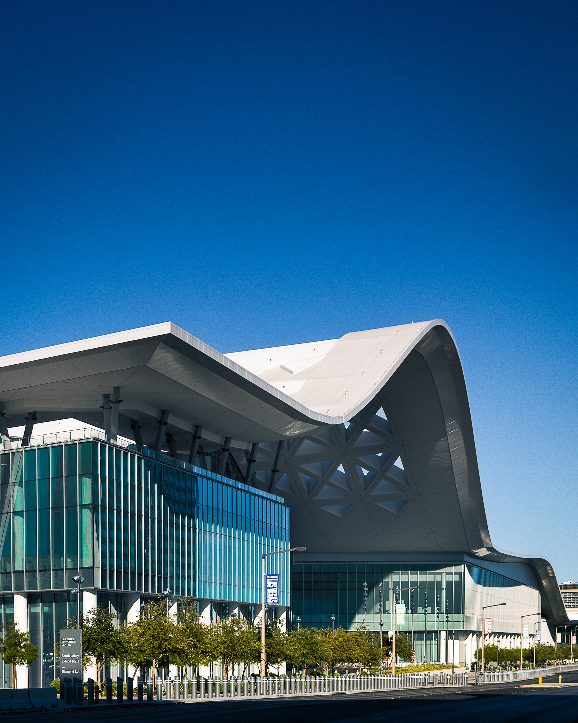 Side view of the Las Vegas Convention Center West Hall showing the sweeping white ribbon roof that echoes the Spring Mountains. The glass-clad meeting-room block sits beneath the cantilevered structure; street-level landscaping and wayfinding signage are in the foreground.