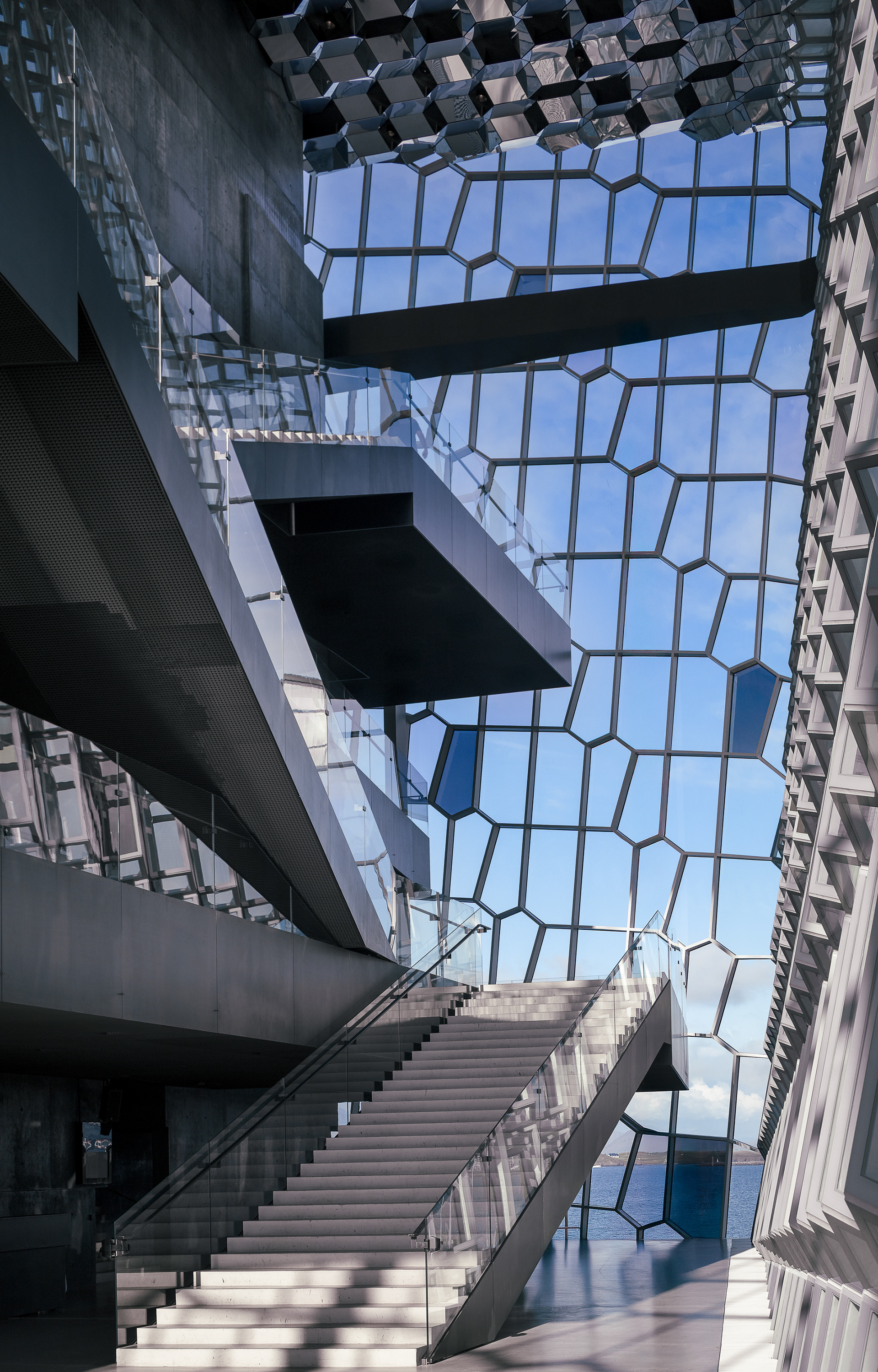 Interior staircase and geometric glass facade inside the Harpa Concert Hall in Reykjavík Iceland with ocean visible through the hexagonal windows.