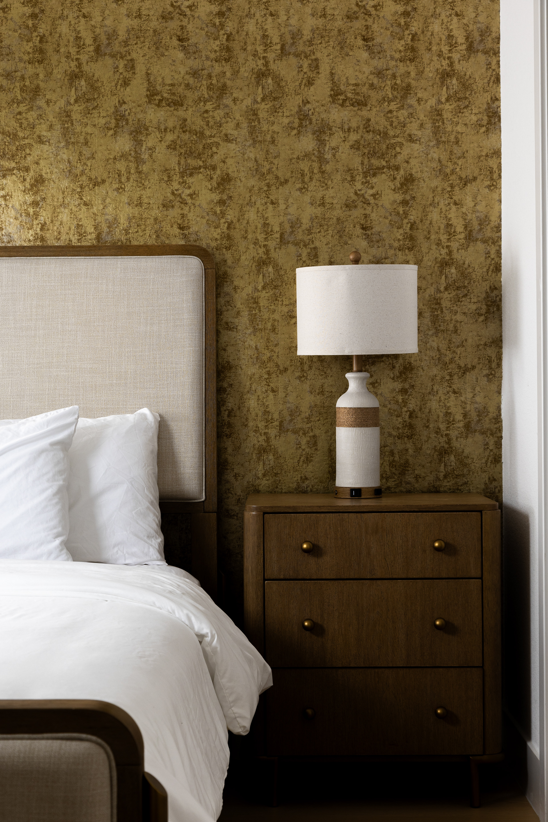 Modern bedroom interior detail in a Las Vegas home featuring a wood nightstand, table lamp, upholstered bed, and textured gold wallpaper.