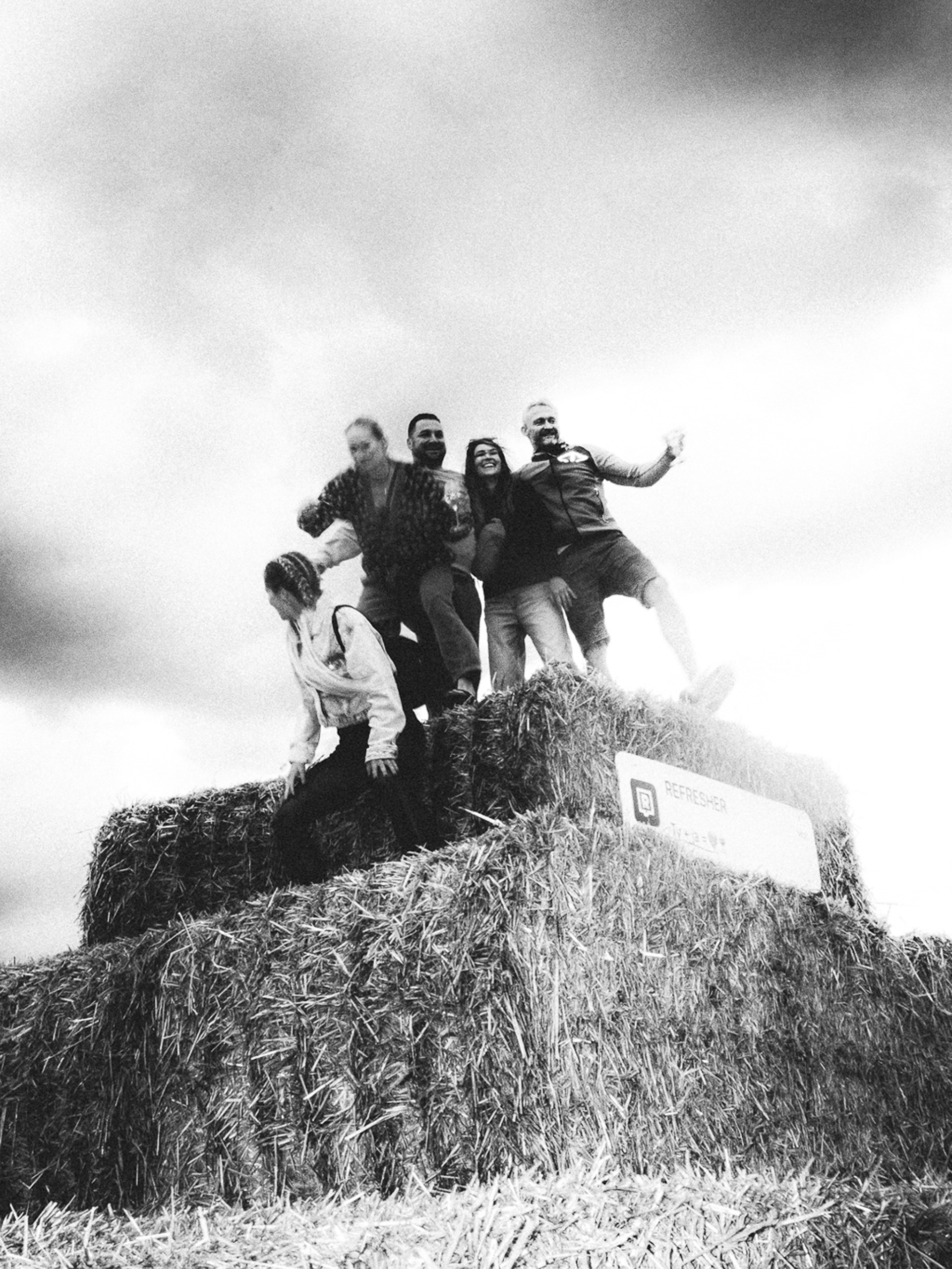Čiernobiela analógová fotografia skupiny ľudí baviacich sa na balíkoch slamy počas festivalu Ženy víno funk 2025.Black and white analog snapshot of a group of people having fun on hay bales at Zeny vino funk festival 2025.