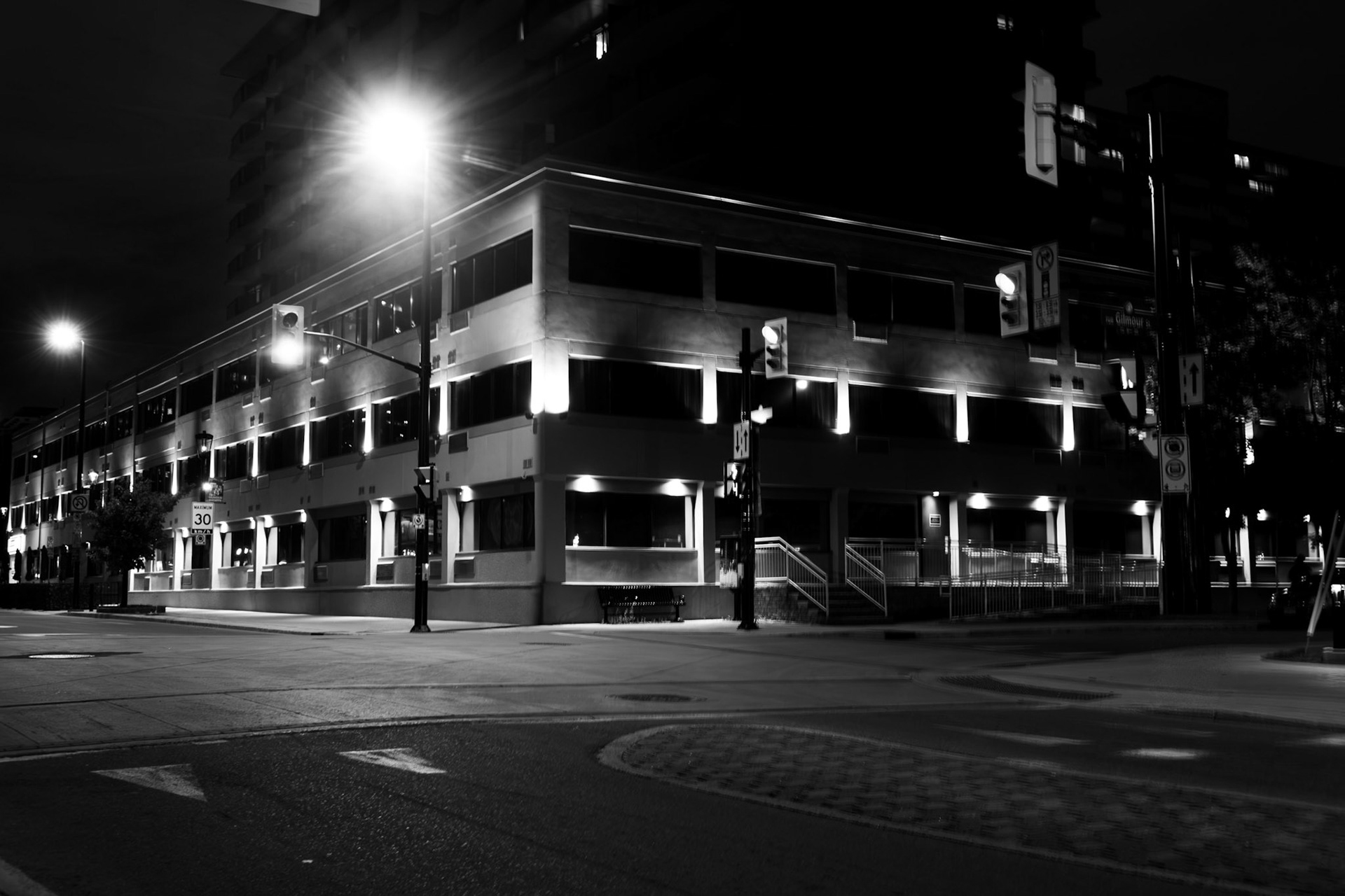 Nocturnal Grid: Monochrome fine art architectural photography capturing a commercial building at night, defined by blazing overhead streetlamps, striking linear window rows, and stark high-contrast shadows.