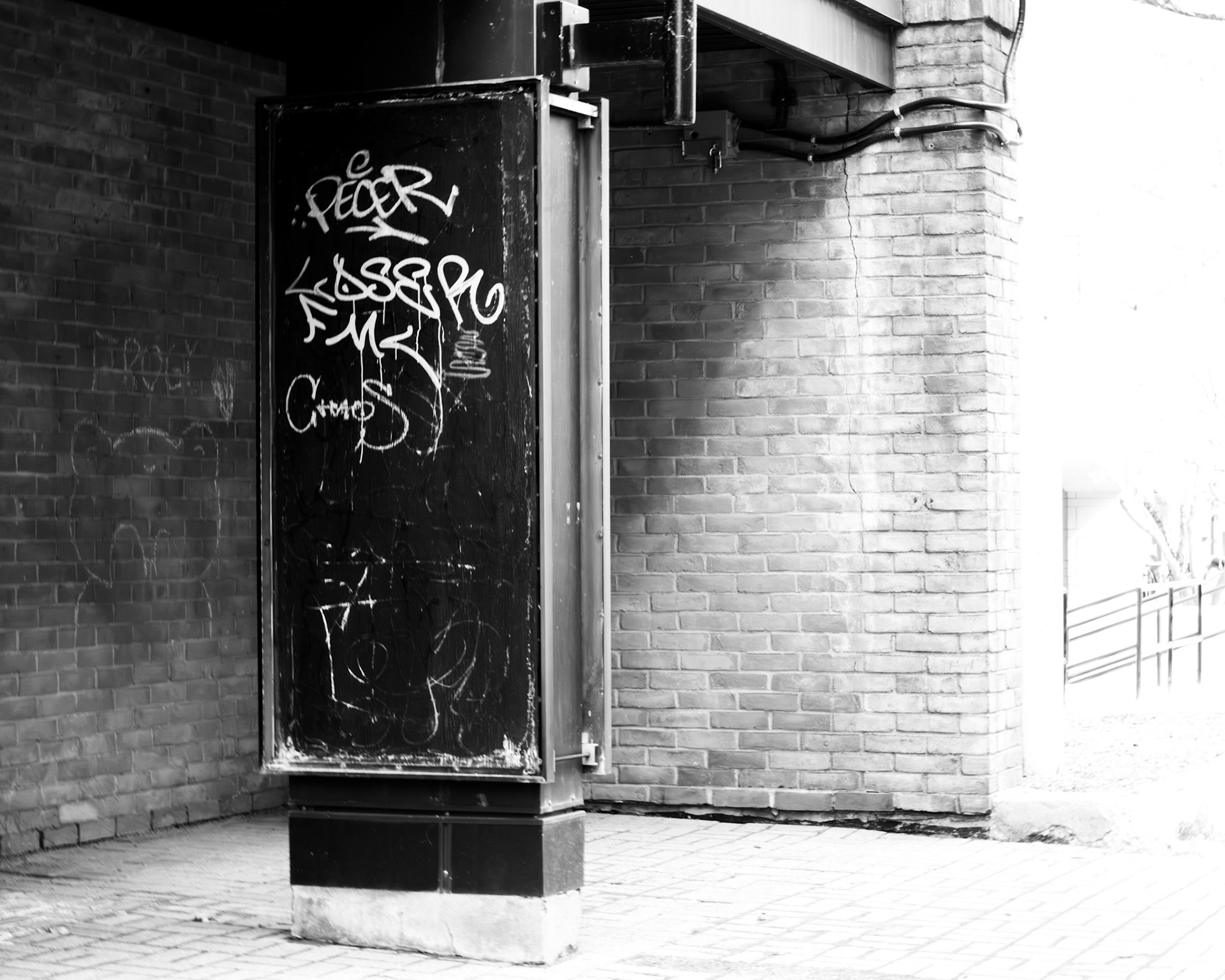 Urban Glyph: Monochrome fine art street photography featuring a dark, graffiti-tagged utility pillar standing in sharp contrast against a weathered brick wall and bright ambient light.