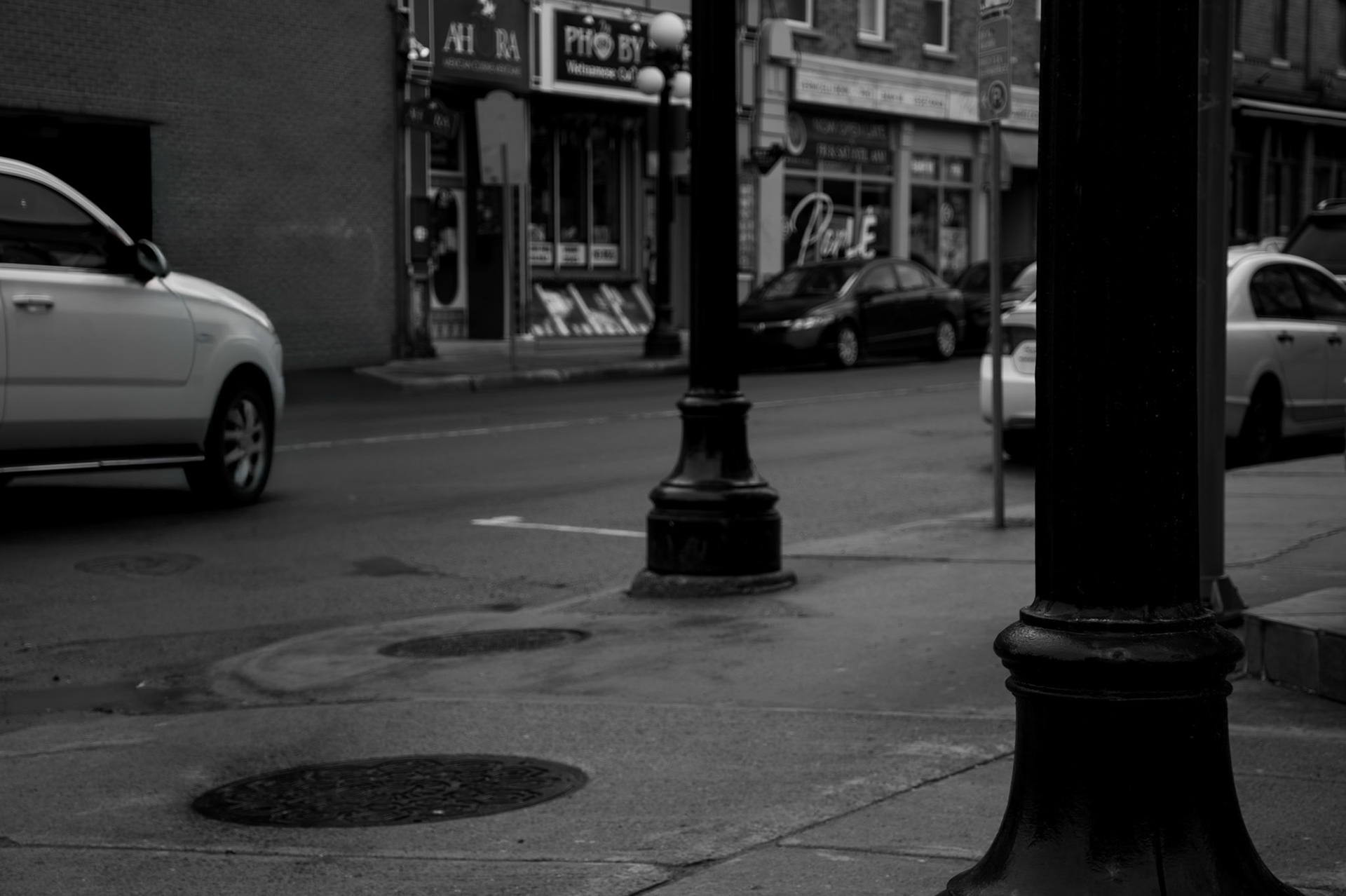 Urban Pillars: Monochrome fine art street photography framing a quiet city street between heavy, dark foreground lamppost columns, featuring parked vehicles, manhole covers, and distant storefronts.