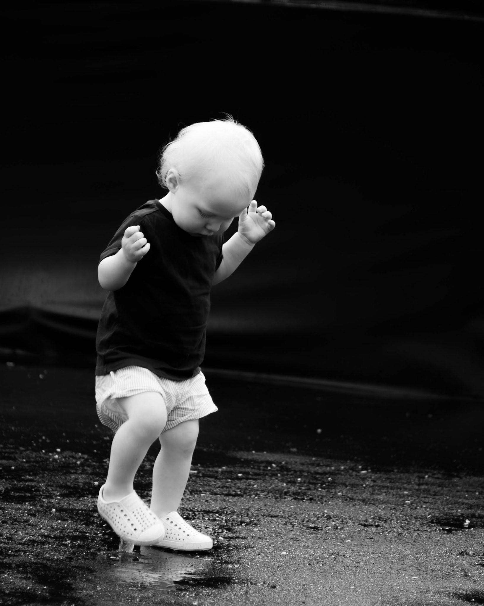 Dancing Child: Monochrome fine art street photography of a toddler with light-colored shoes dancing, observing his reflection on a wet surface against a deep, dark background, capturing a moment of innocent movement and isolation.